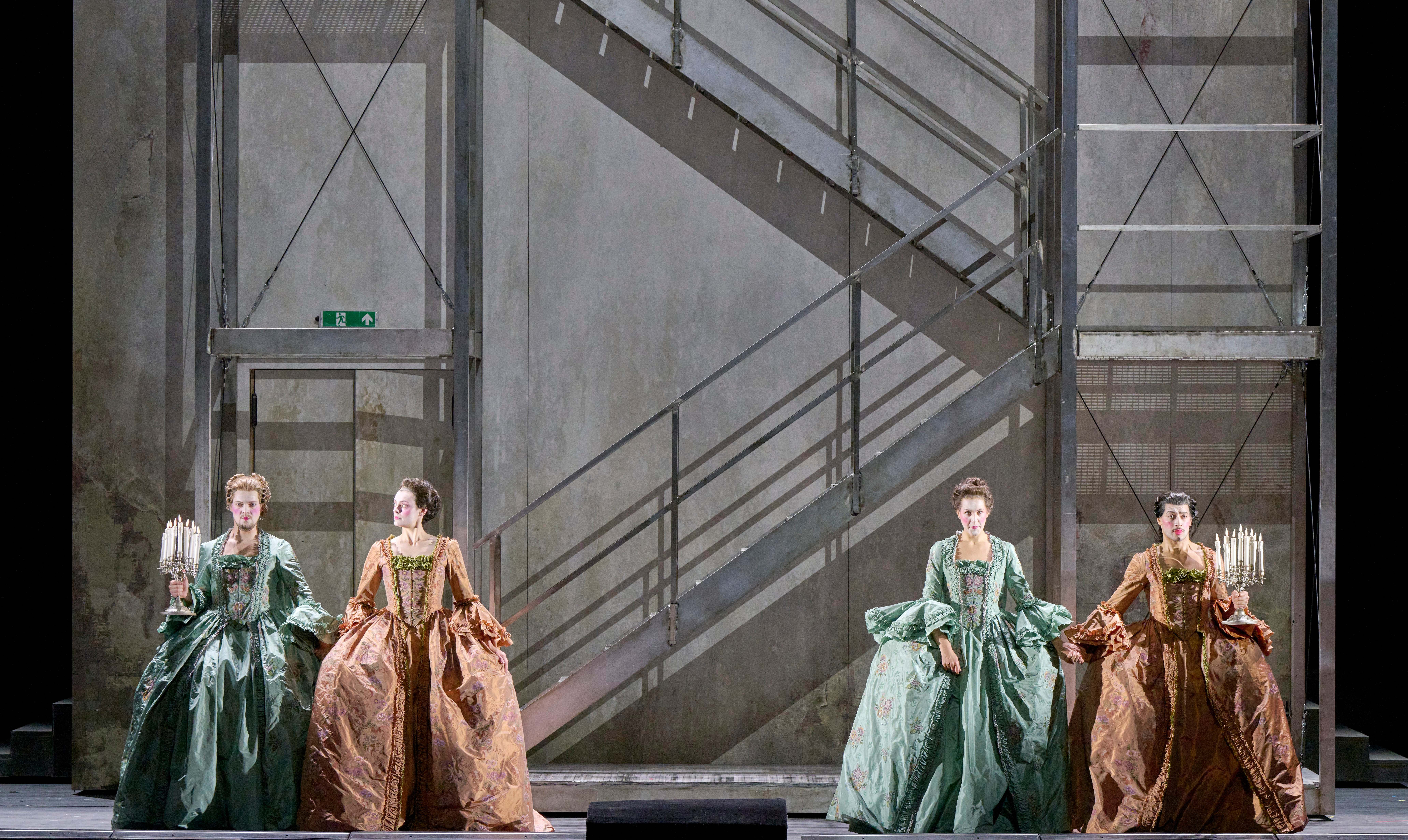 Vier Frauen in prunkvollen barocken Kleidern stehen nebeneinander auf einer Bühne vor einer nüchternen, grauen Wand mit diagonalen Metalltreppen.