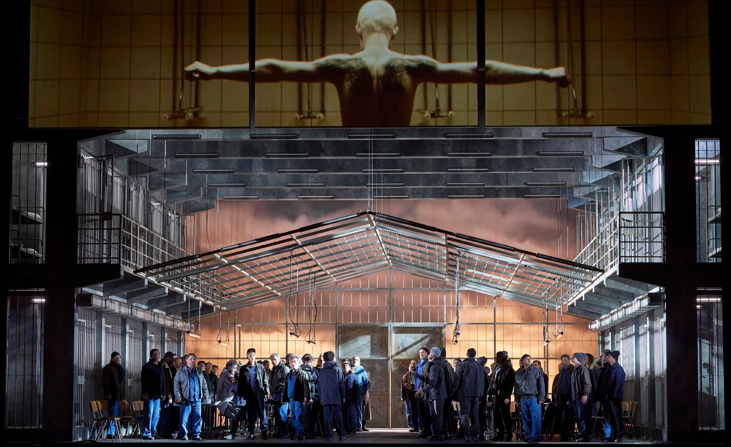 Vor einem transparenten Gebäude versammelt sich eine große Gruppe Menschen, darüber ist ein Bild eines nackten, ausgebreiteten Oberkörpers zu sehen.