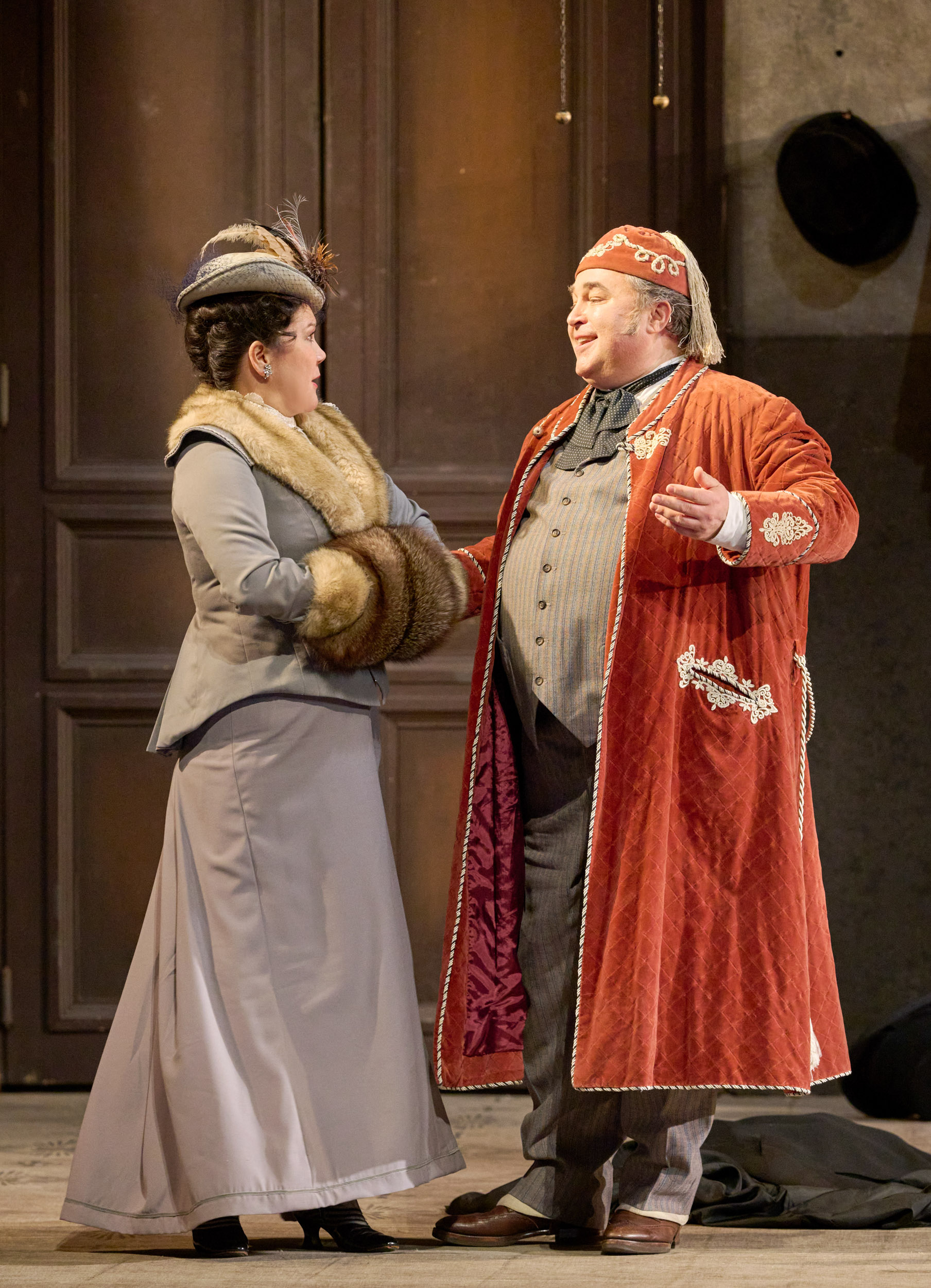 Eine elegant gekleidete Frau mit Pelzstola steht einem Mann im roten Morgenmantel gegenüber, der mit offenen Armen spricht.