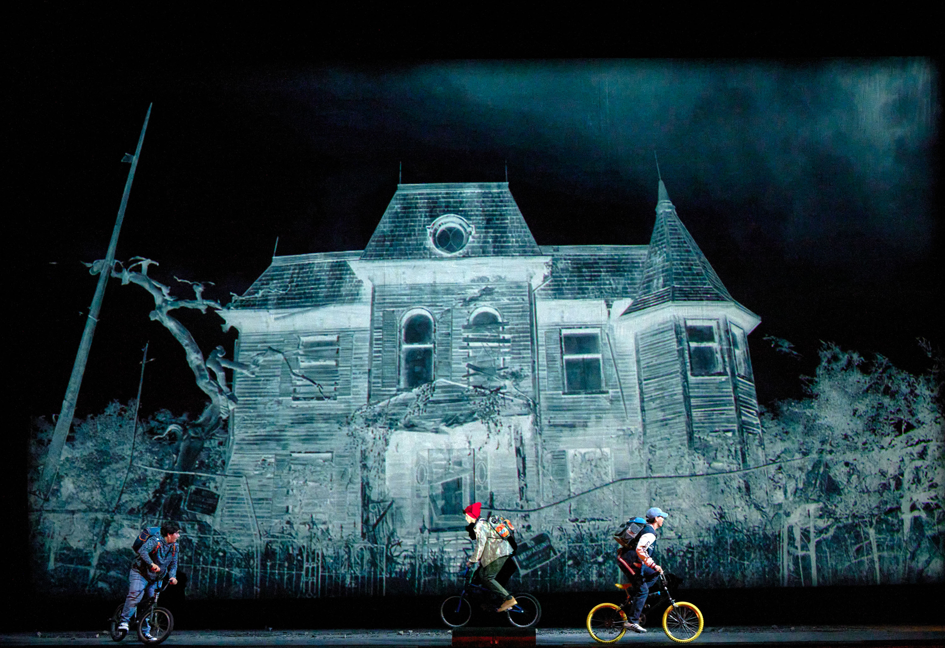 Drei Kinder fahren auf kleinen Fahrrädern vor der Projektion eines unheimlich wirkenden alten Hauses.