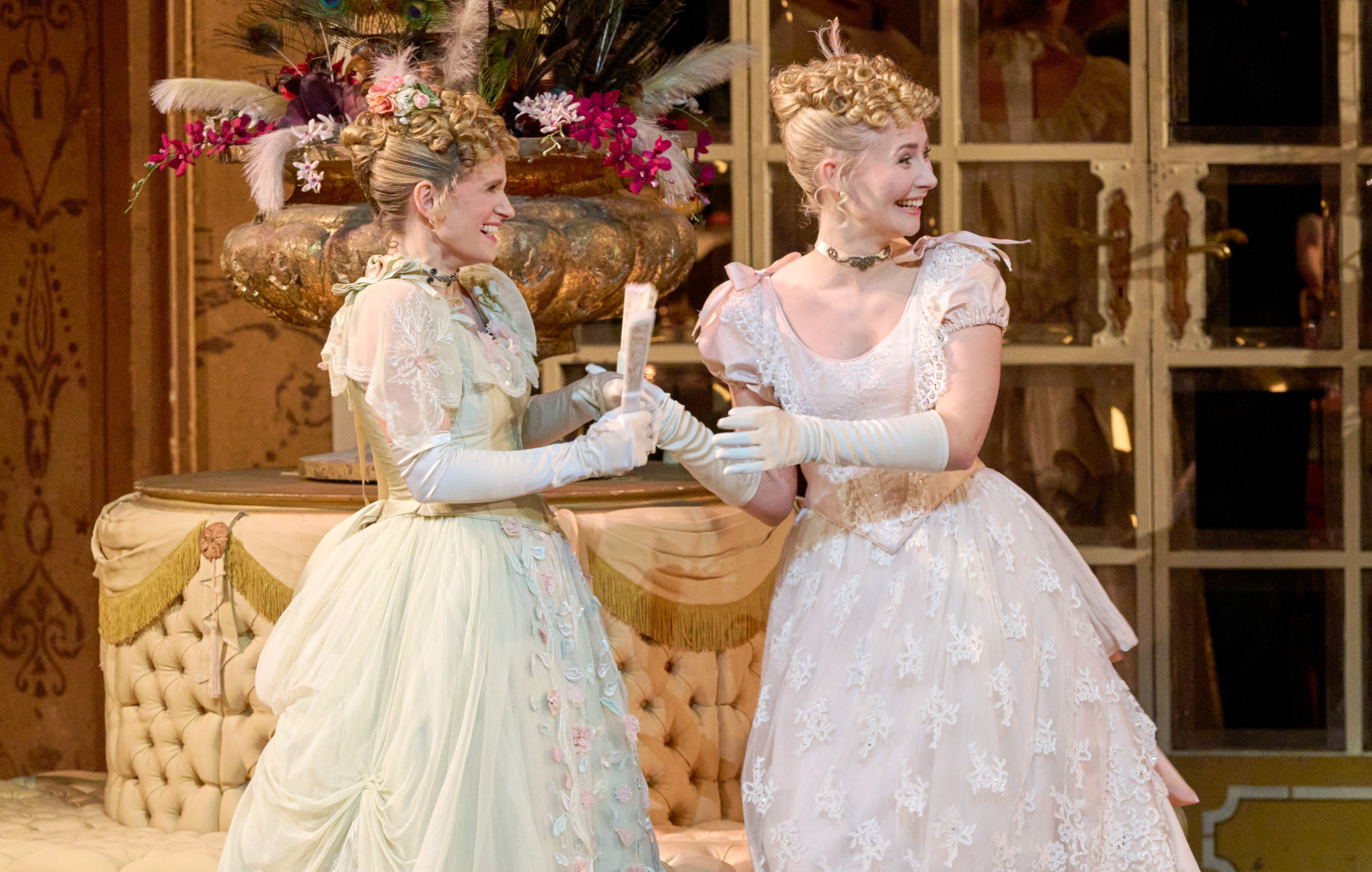 Zwei junge Frauen in prachtvollen Ballkleidern lachen ausgelassen und halten gemeinsam eine Schriftrolle in festlicher Umgebung.