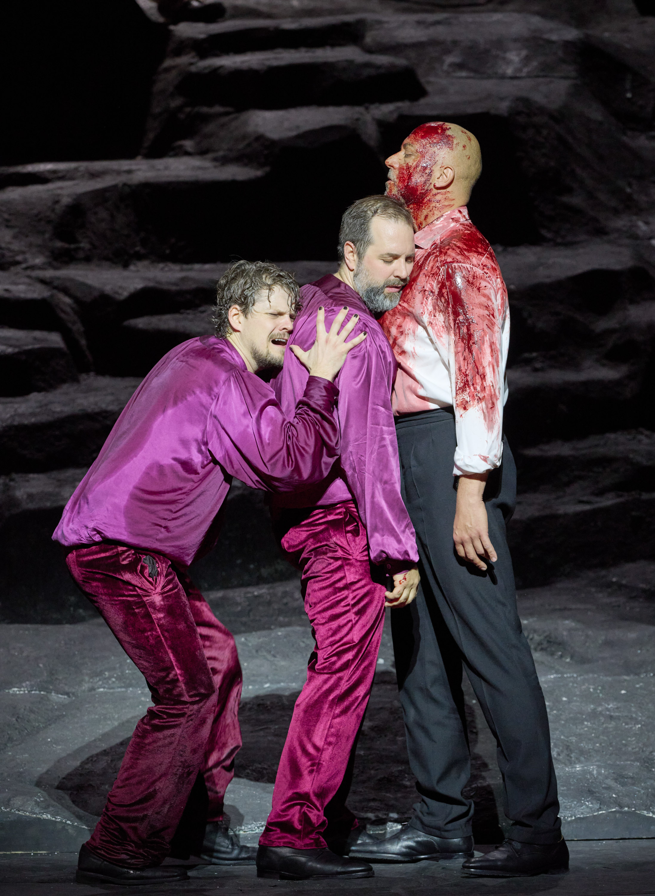 Zwei Männer in einem roten Samt-Gewand lehnen sich an einen blutigen dritten Mann, der aufrecht vor ihnen steht.