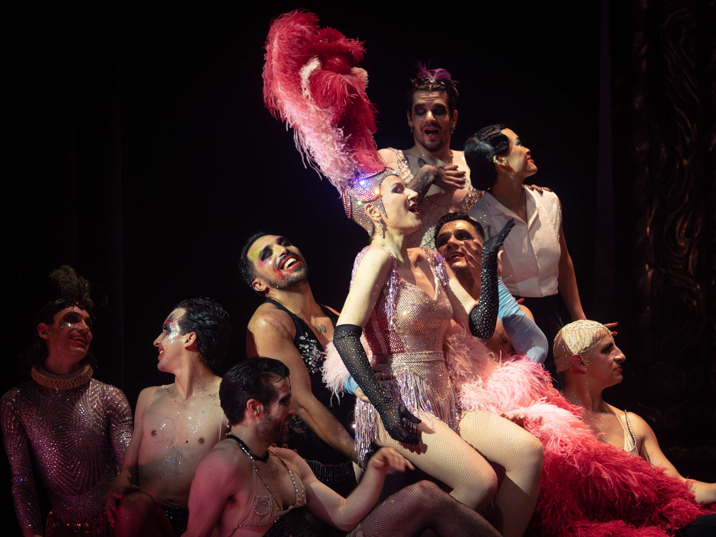 Eine Frau in einem pinken Glitzerkostüm und einer Federkrone auf dem Kopf sitzt auf einem Haufen Männern