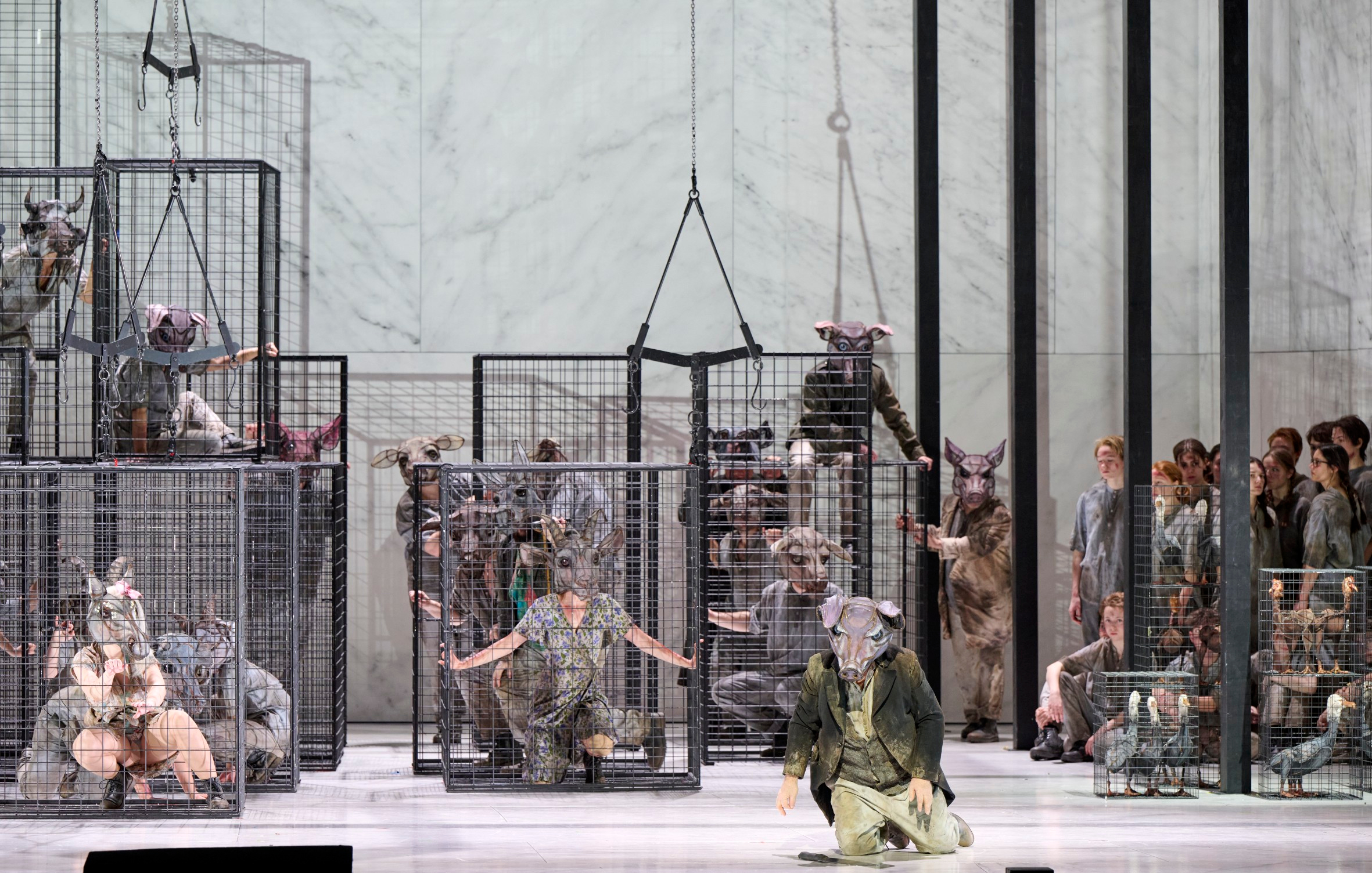 Mehrere Personen in Tiermasken befinden sich in und um großen Käfigen, während ein künstlich beleuchteter, moderner Raum sie umgibt.