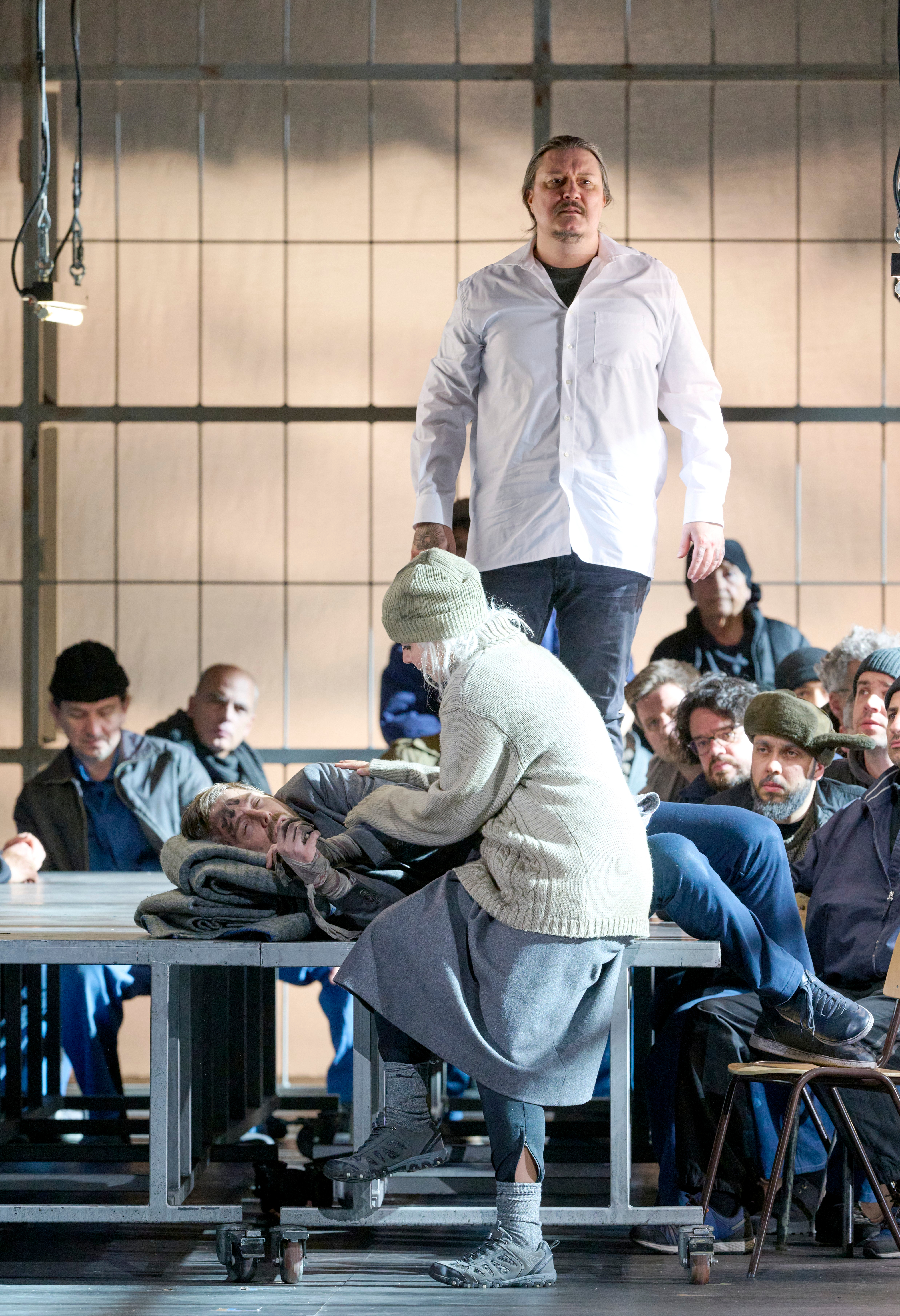 Ein Mann in weißem Kittel steht erhoben vor einer sitzenden Menschenmenge, während eine Person im Vordergrund medizinische Hilfe leistet.