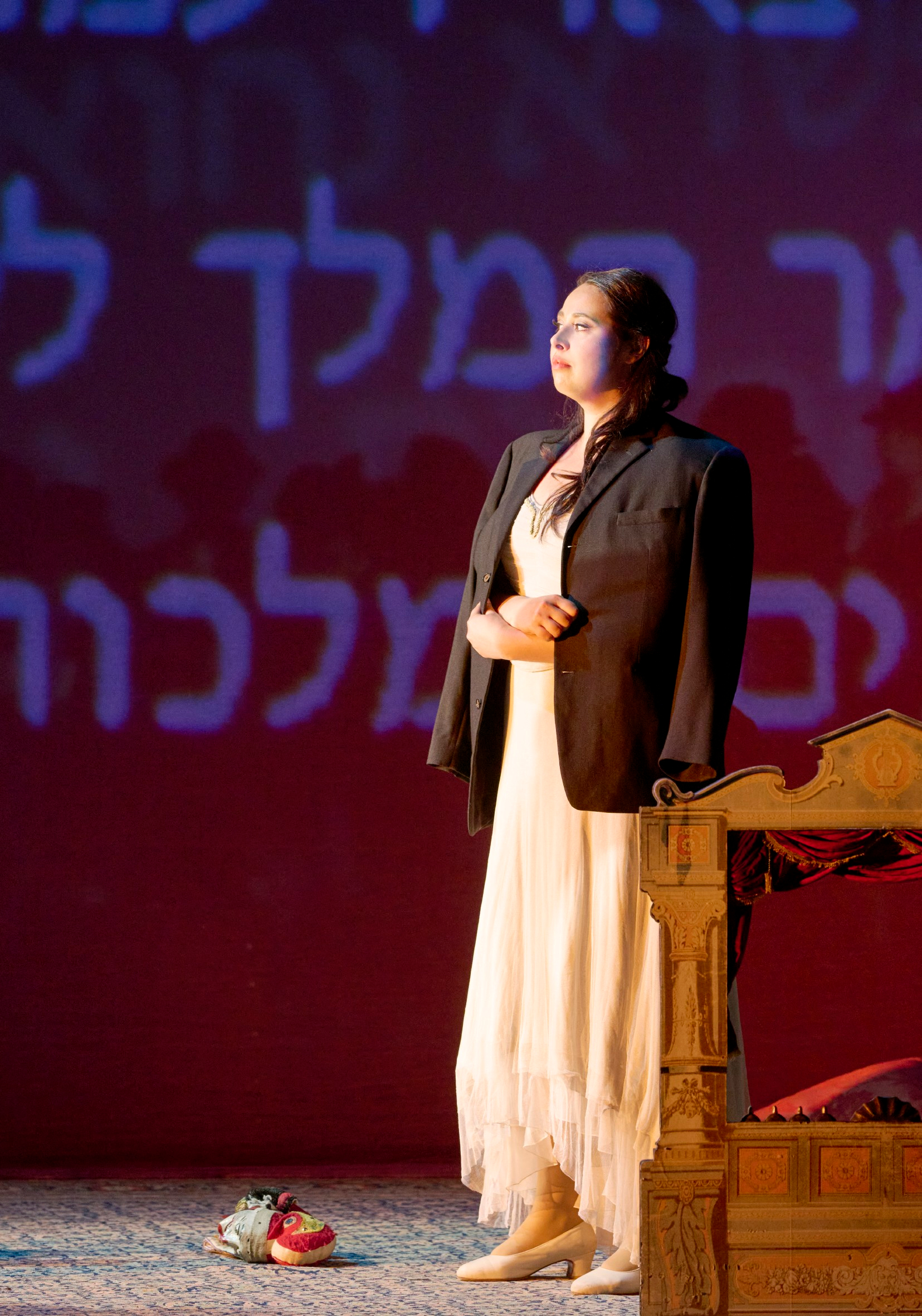 Eine Frau in hellem Kleid und dunklem Jackett steht vor hebräischer Schriftprojektion auf rotem Bühnenhintergrund.