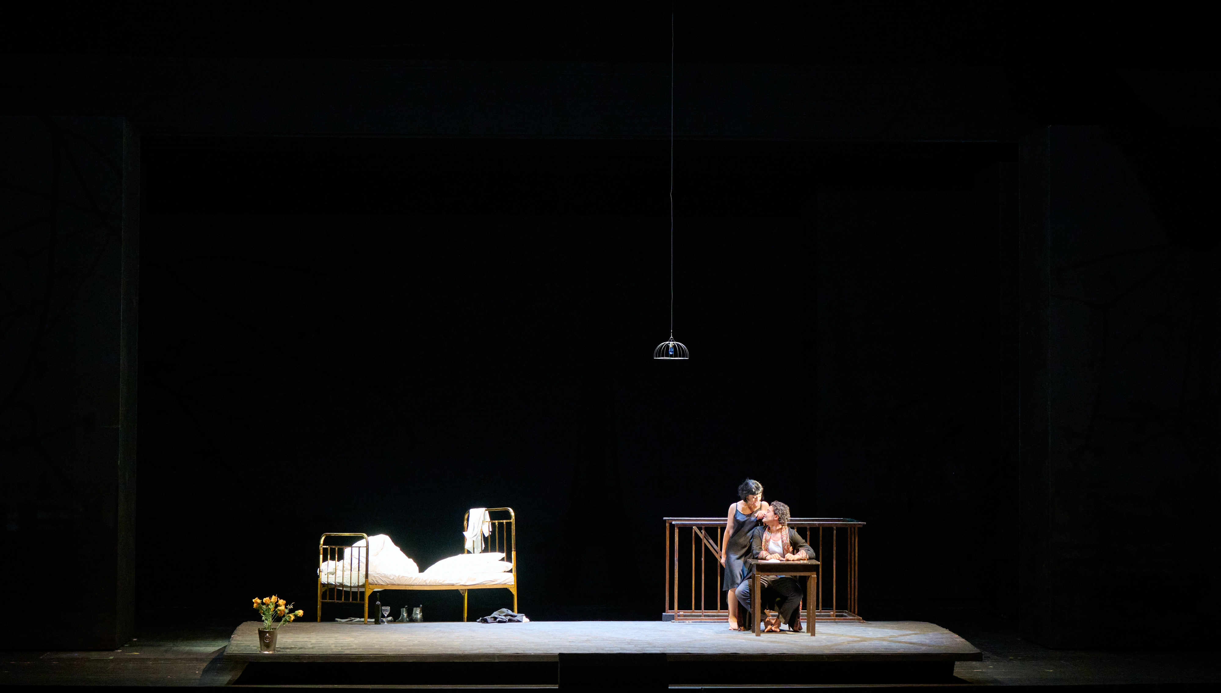 Zwei Personen sitzen eng umschlungen an einem Tisch auf einer spärlich beleuchteten Bühne; links steht ein leeres Bett unter einer Hängelampe.