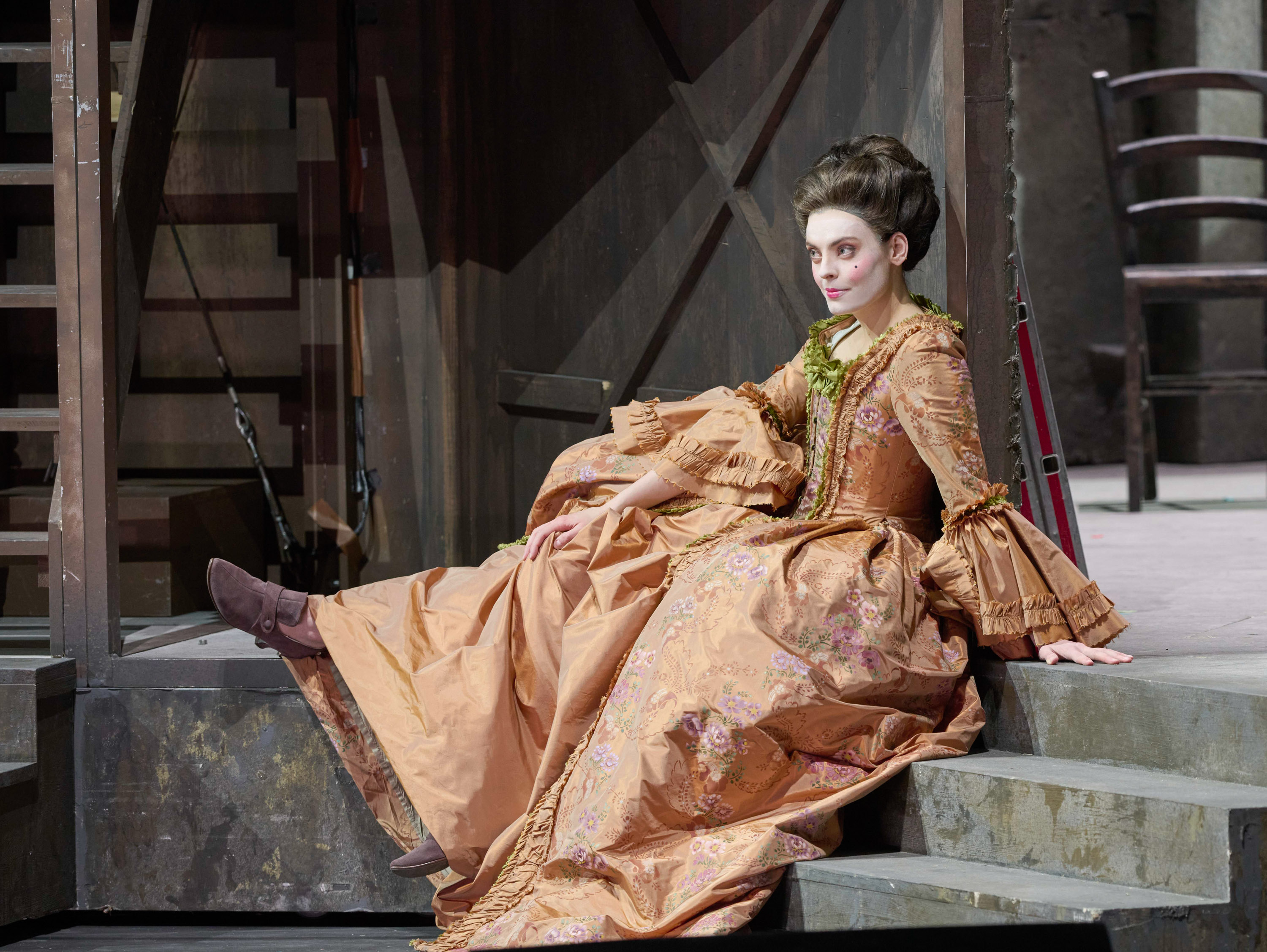 Eine Frau in einem aufwendig verzierten, kupferfarbenen Rokoko-Kleid sitzt mit leicht geneigtem Kopf auf einer Treppe und blickt nachdenklich zur Seite.