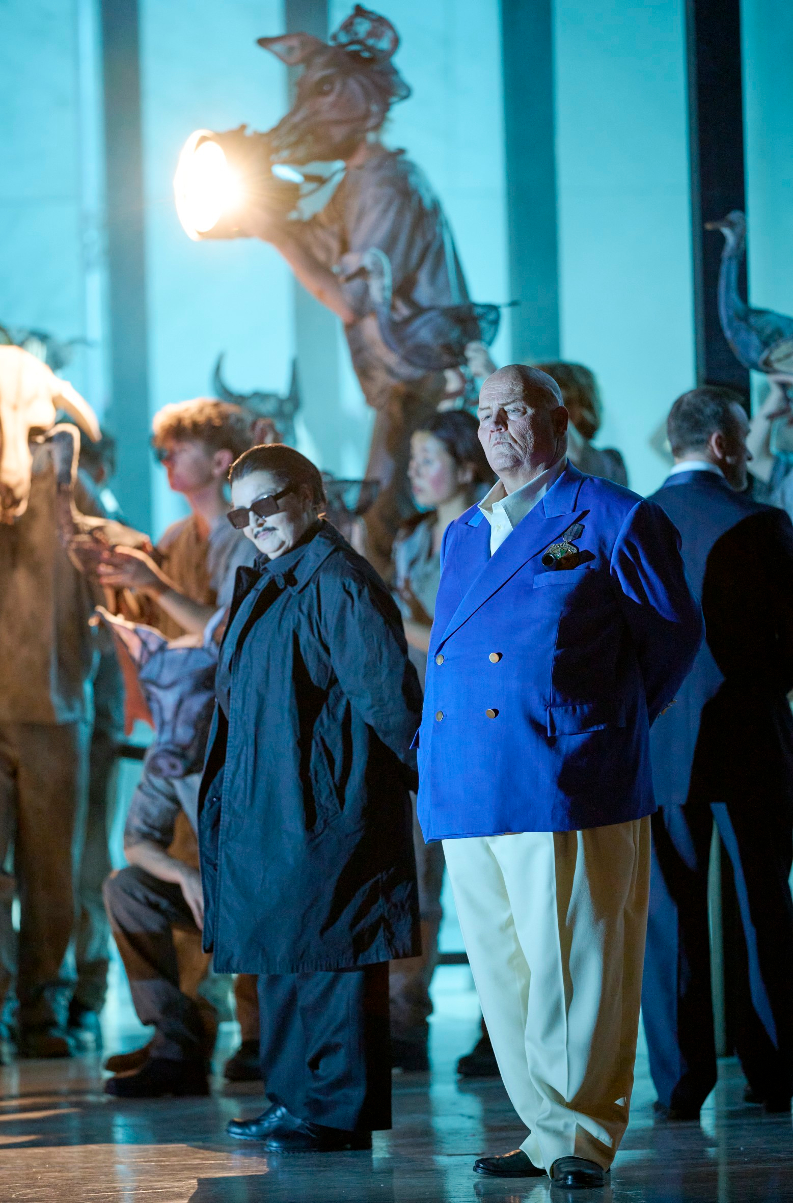 Zwei Männer, einer in dunkler Sonnenbrille und der andere in blauem Sakko, stehen vor einer skurrilen Szene mit Tiermasken und grellem Licht.