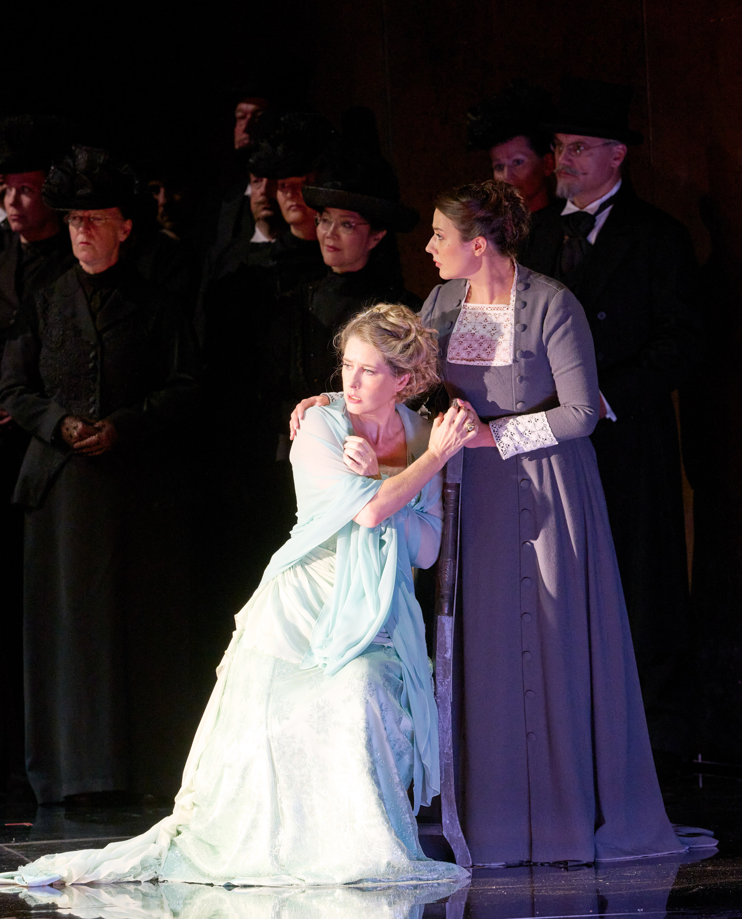 Zwei Frauen, die eine in einem hellen und die andere in einem dunklen Kleid, stehen vor einer Gruppe schwarz gekleideter Menschen.