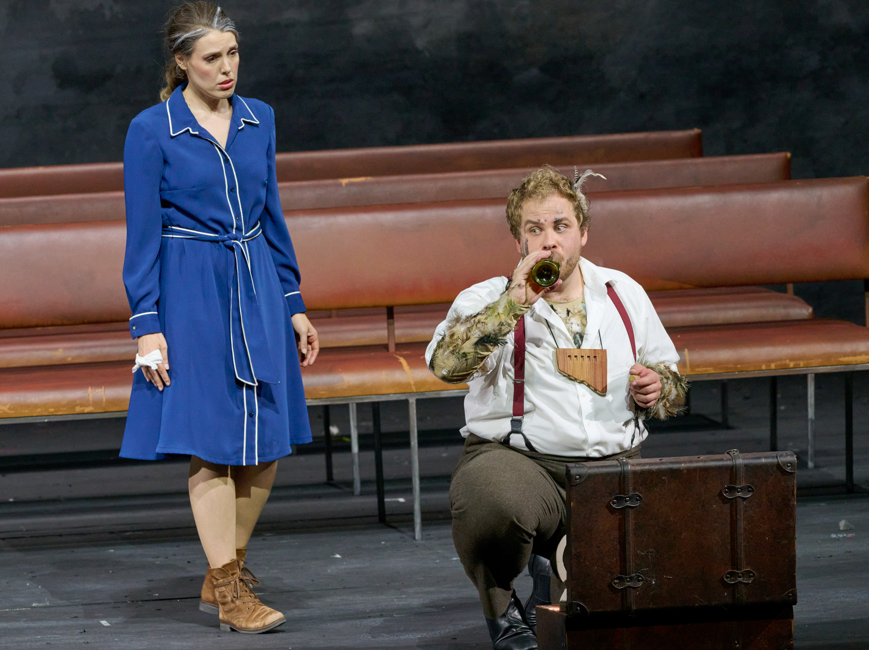 Ein Mann mit Federn in den Haaren und einer Panflöte um den Hals kniet vor einem Koffer und trinkt aus einer Glasflasche. Hinter ihm steht eine junge Frau in einem blauen Kleid und betrachtet ihn besorgt.