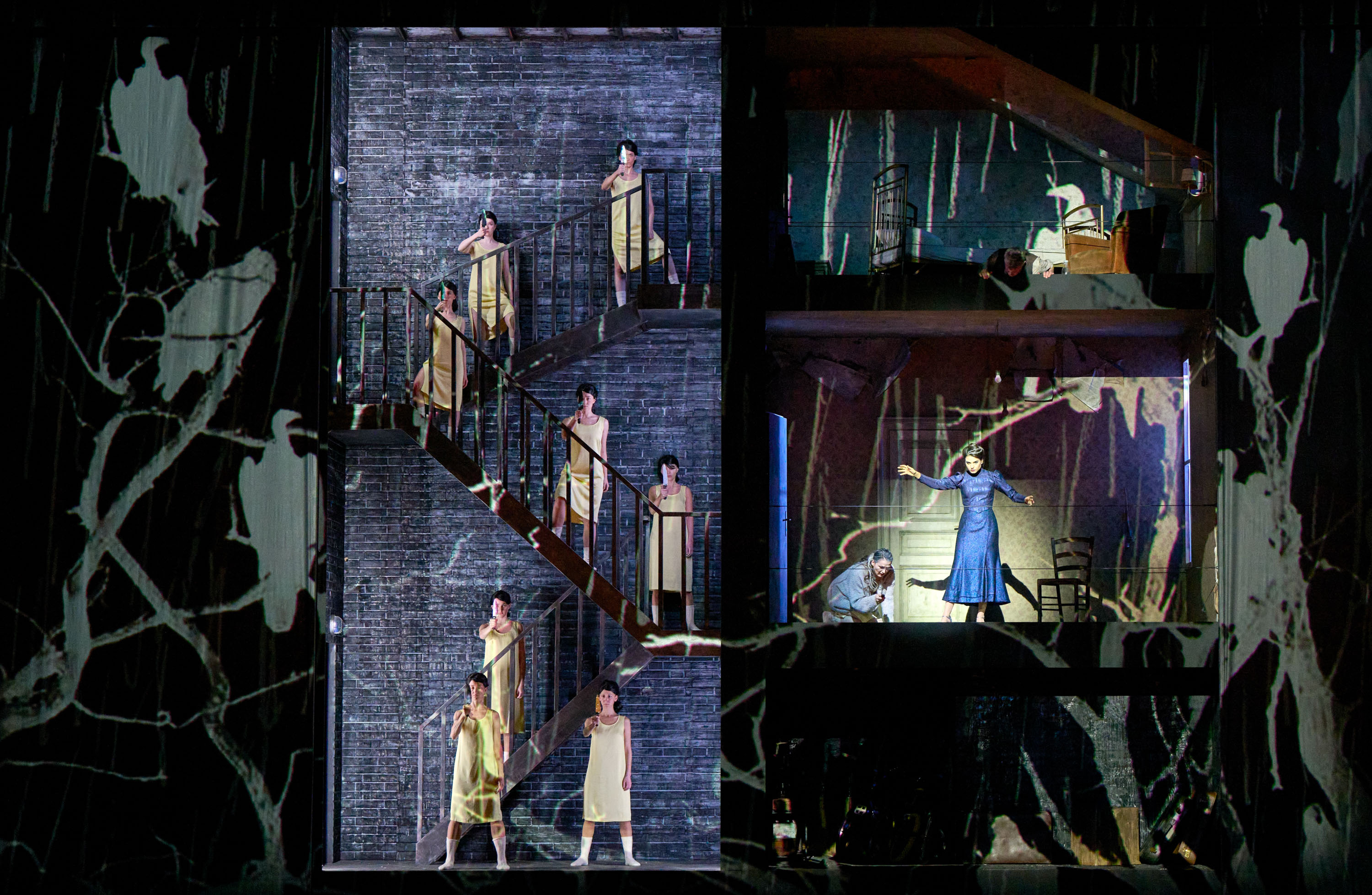 Auf einer Bühne sind mehrere Stockwerke eines Hauses abgebildet, mit Darstellern in weißen Kleidern, die in verschiedenen Posen auf Treppen und in Räumen stehen.