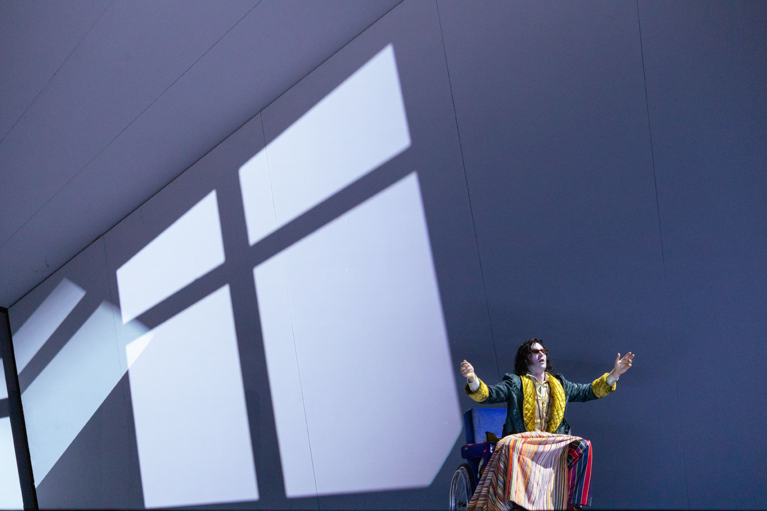 Eine Person in farbenfrohem Kostüm sitzt mit ausgebreiteten Armen an einem Tisch vor einer großen, schräg beleuchteten Wand.