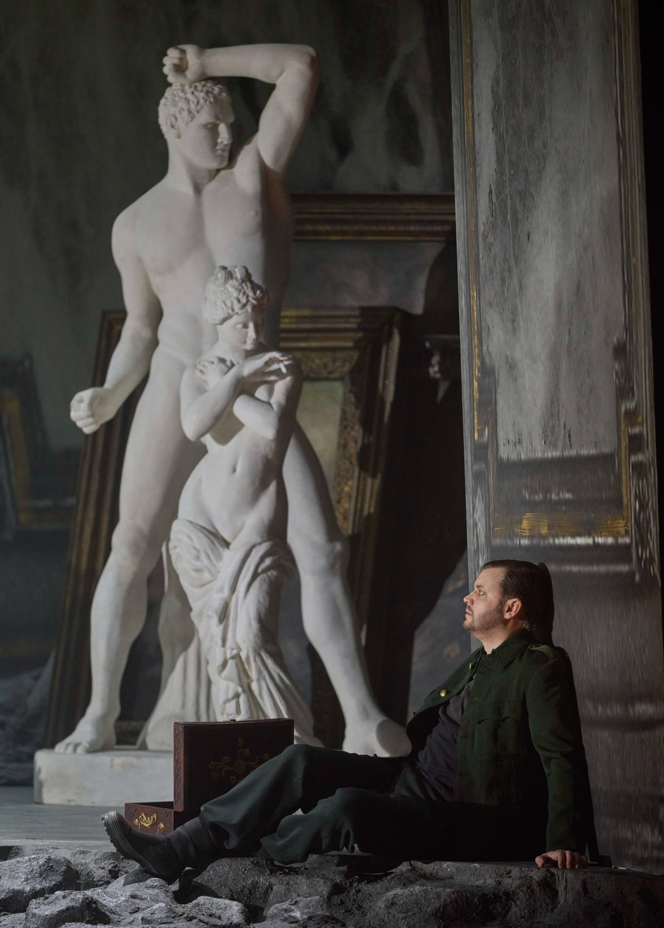 Ein Mann in dunkler Kleidung sitzt nachdenklich vor einer weißen Marmorskulptur und einer geöffneten Truhe auf steinigem Boden.
