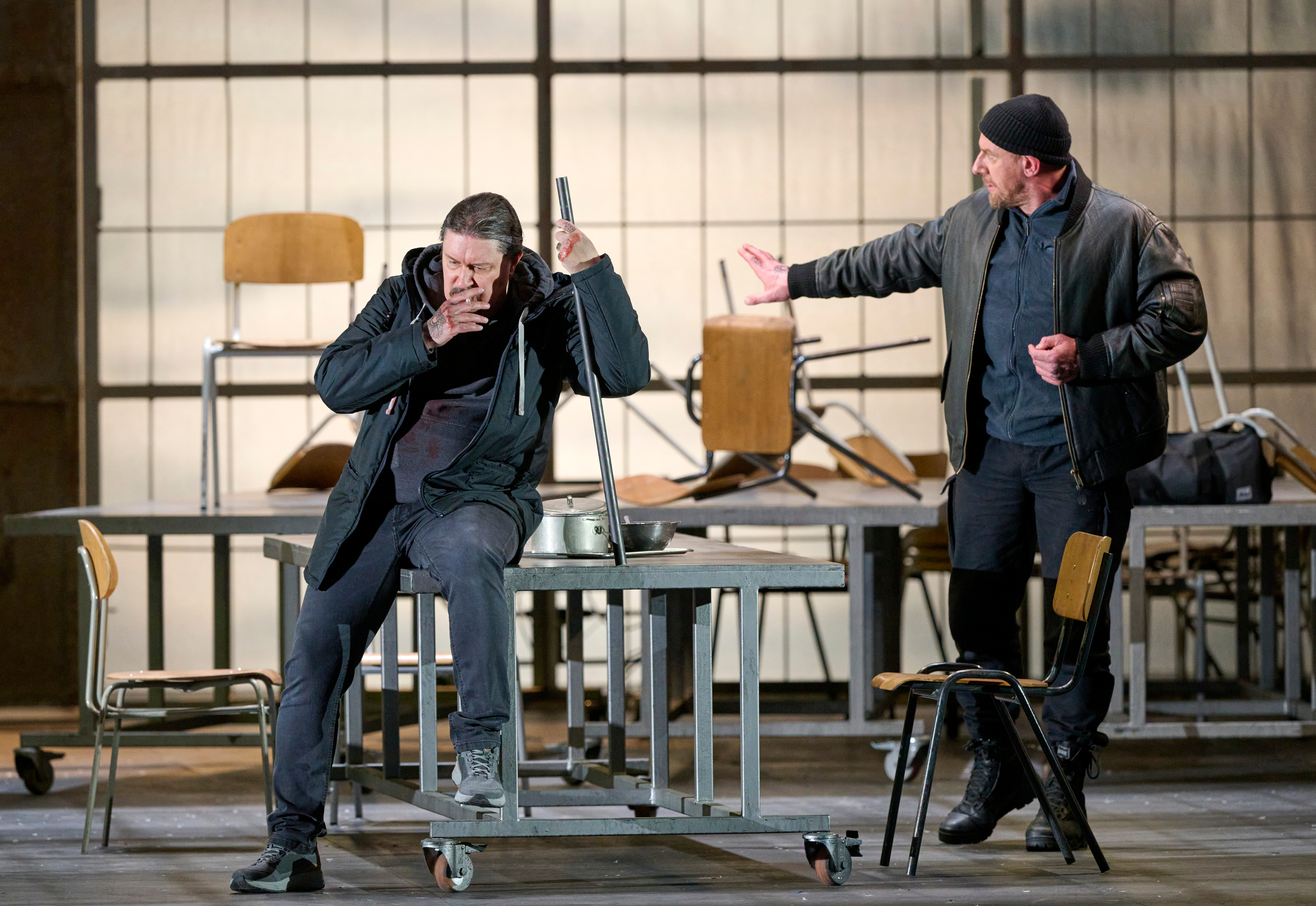 Zwei Männer befinden sich in einem Raum mit umgestürzten Tischen; der eine sitzt erschöpft auf einem Stuhl, der andere gestikuliert energisch.