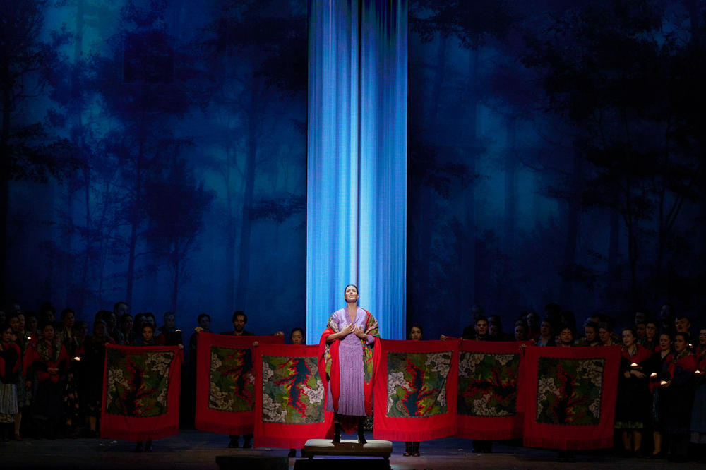 Eine Person im prachtvollen Gewand steht erhöht vor einem Chor, der rote Tücher mit bunten Motiven hält, auf einer Bühne mit Waldkulisse.