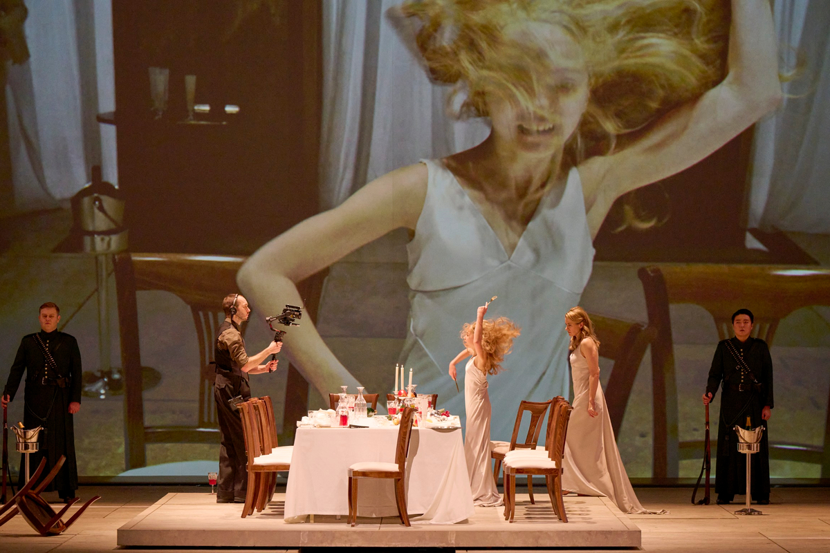Drei Personen stehen um einen festlich gedeckten Tisch, während im Hintergrund eine riesige Projektion einer tanzenden Frau in weißem Kleid zu sehen ist.