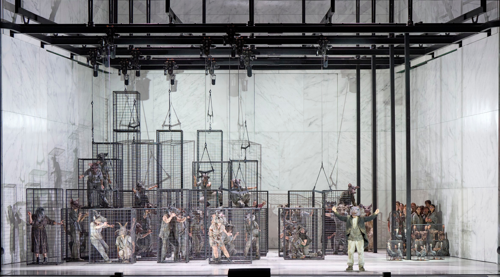 Eine Person mit ausgebreiteten Armen steht vor mehreren Käfigen, in denen Menschen mit Tiermasken eingesperrt sind; der Hintergrund ist kahl und weiß.
