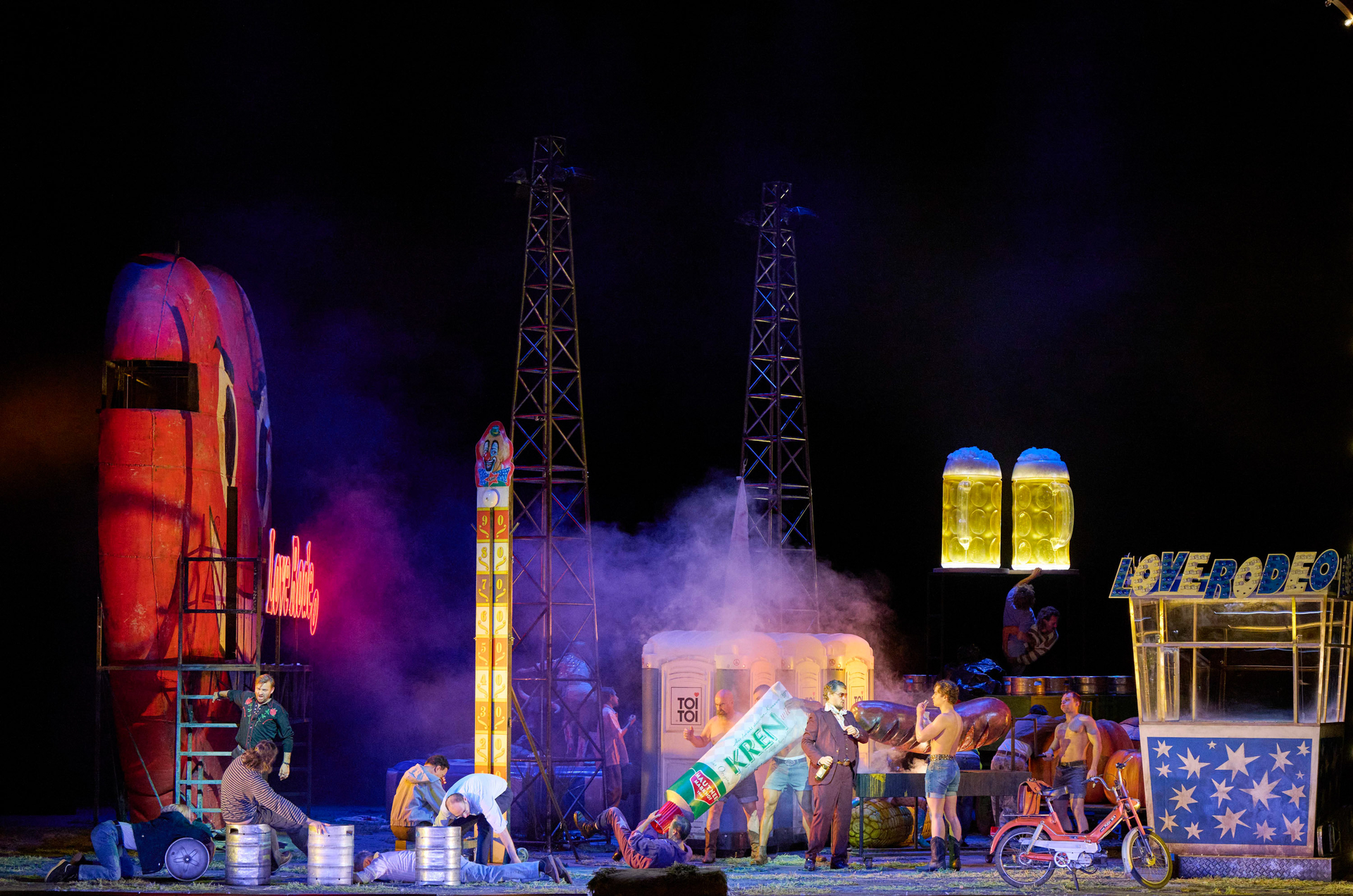 Viele Menschen befinden sich in einer chaotischen Kirmes-Kulisse. Während manche bei einer großen Kren-Tube und einer riesigen Bratwurst sind, liegen und sitzen andere um Metallfässer herum.