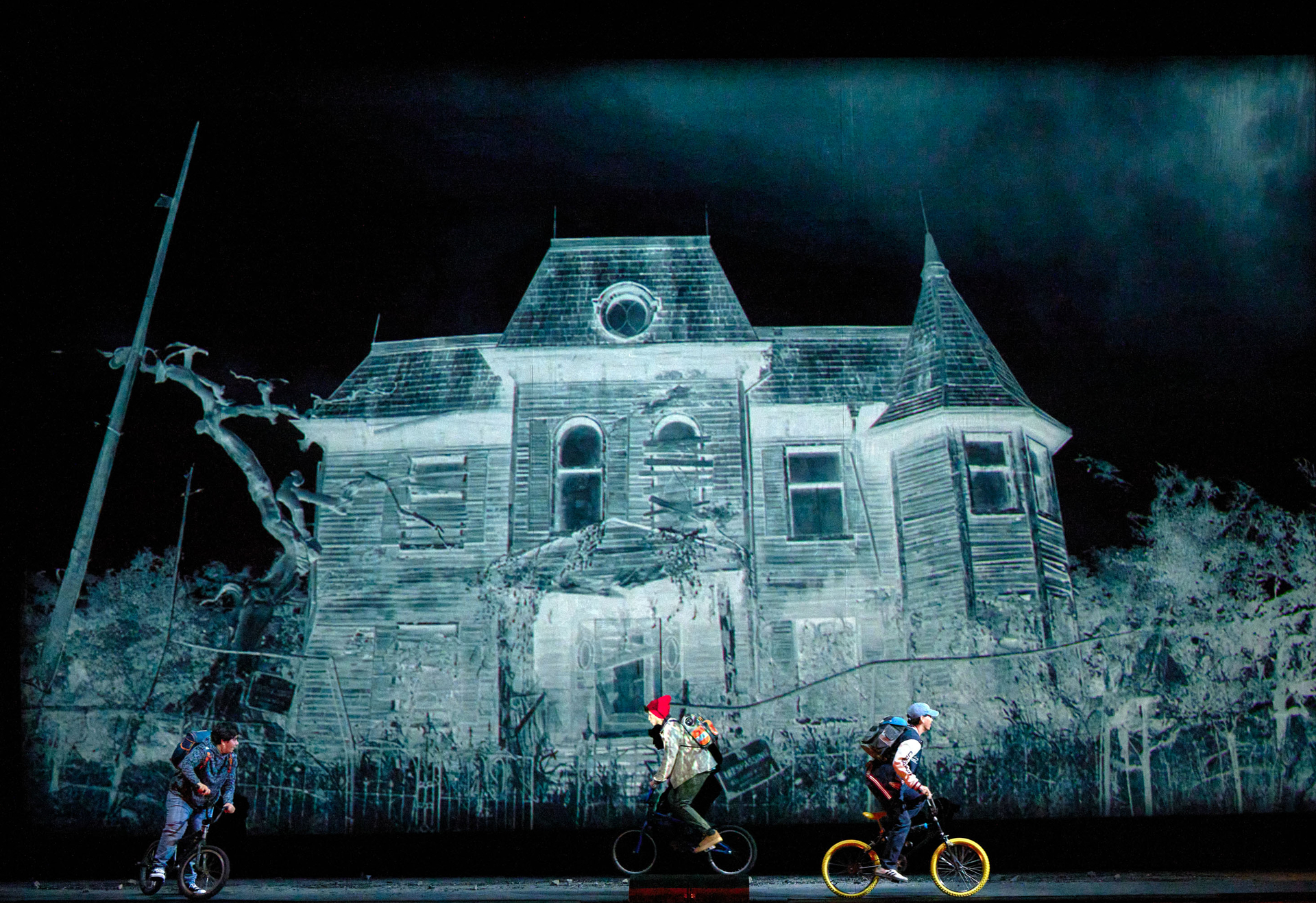 Drei Kinder fahren auf kleinen Fahrrädern vor der Projektion eines unheimlich wirkenden alten Hauses.