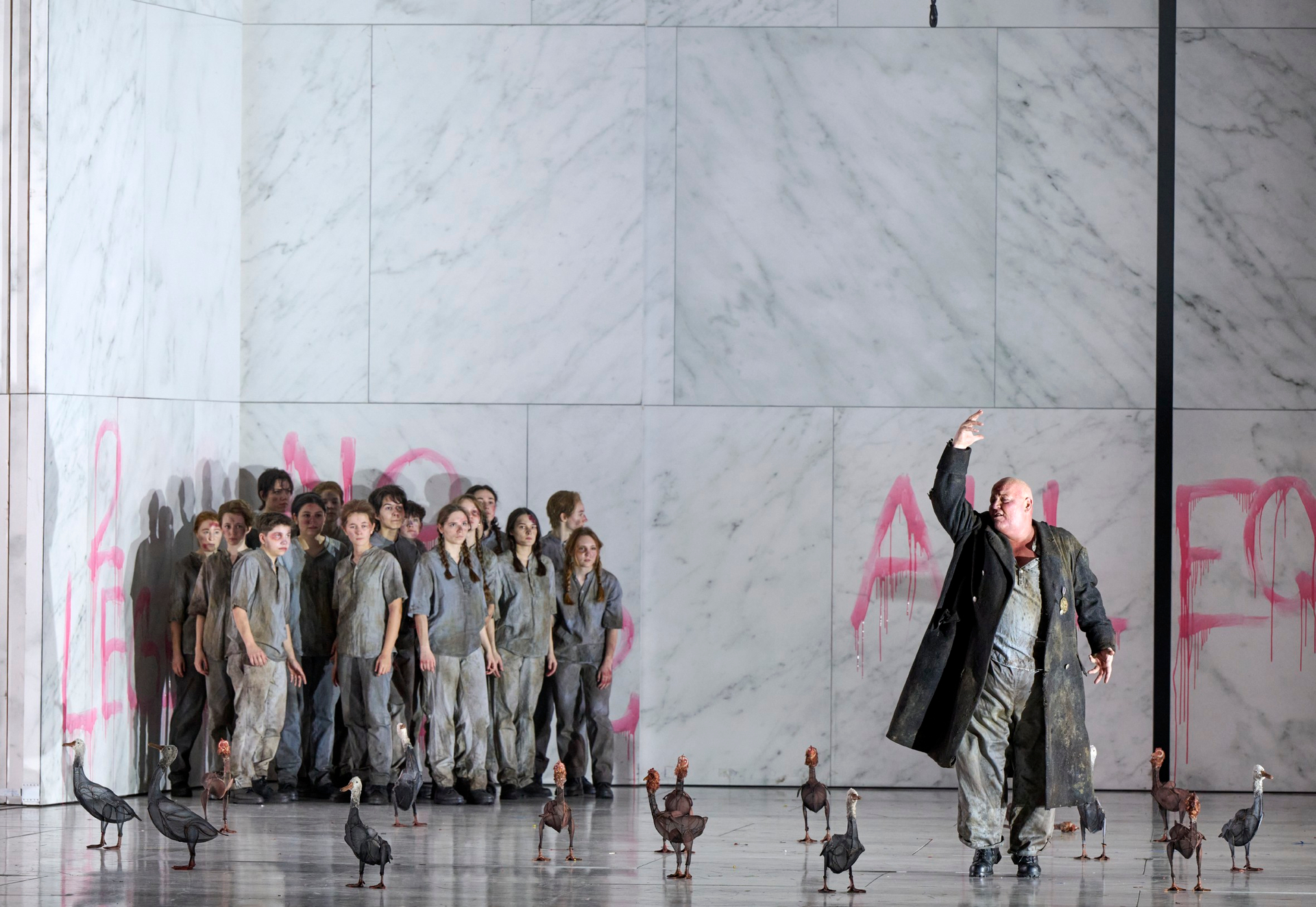 Ein Mann in langem Mantel gestikuliert auf einer Opernbühne mit Vögeln, während eine Gruppe in grauen Overalls vor einer Marmorwand mit rotem Graffiti steht.