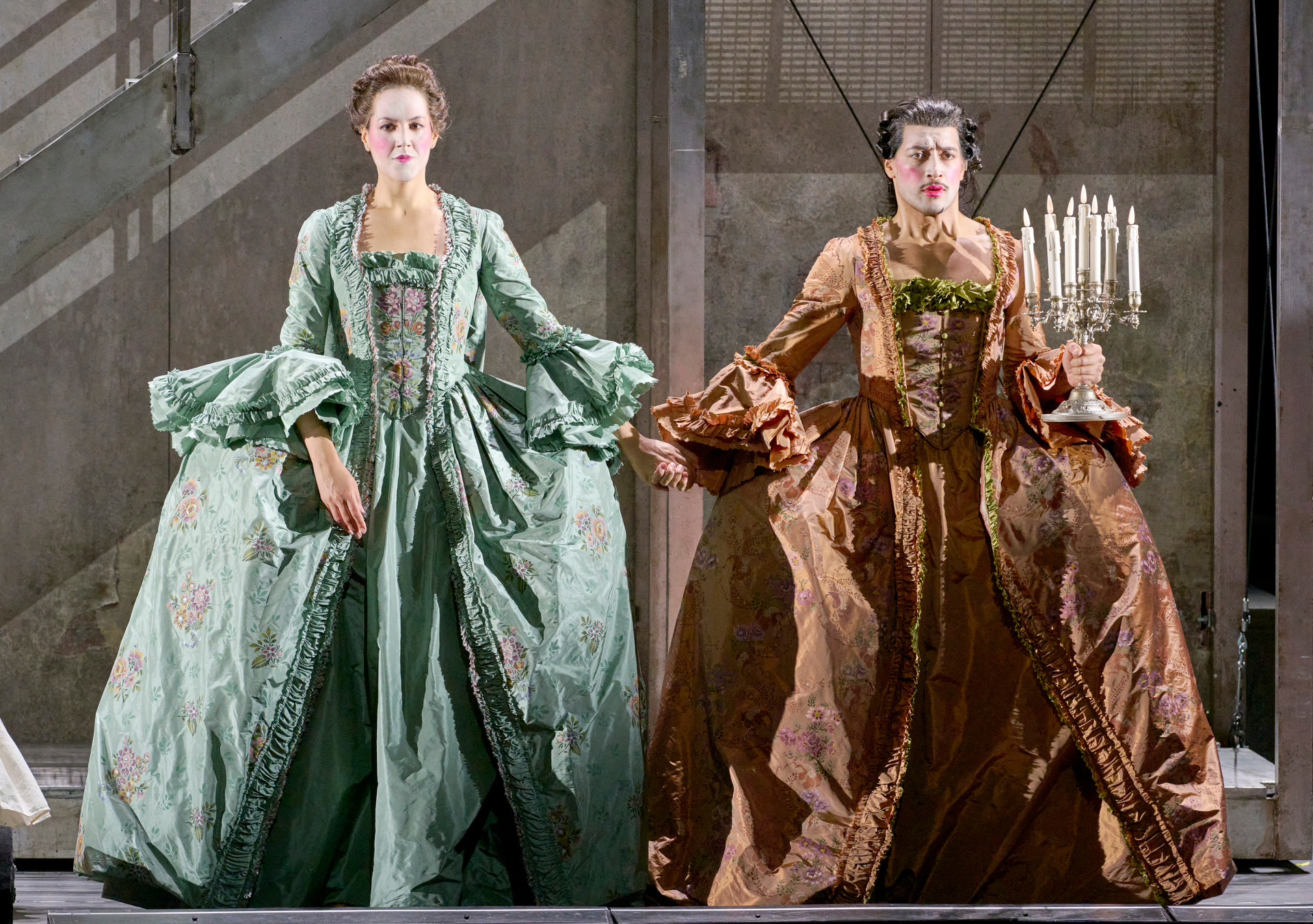 Zwei Frauen in prächtigen barocken Kleidern – eine in Grün, die andere in Kupfer – stehen nebeneinander auf der Bühne, beide mit ernstem Blick und ausgestreckten Armen.