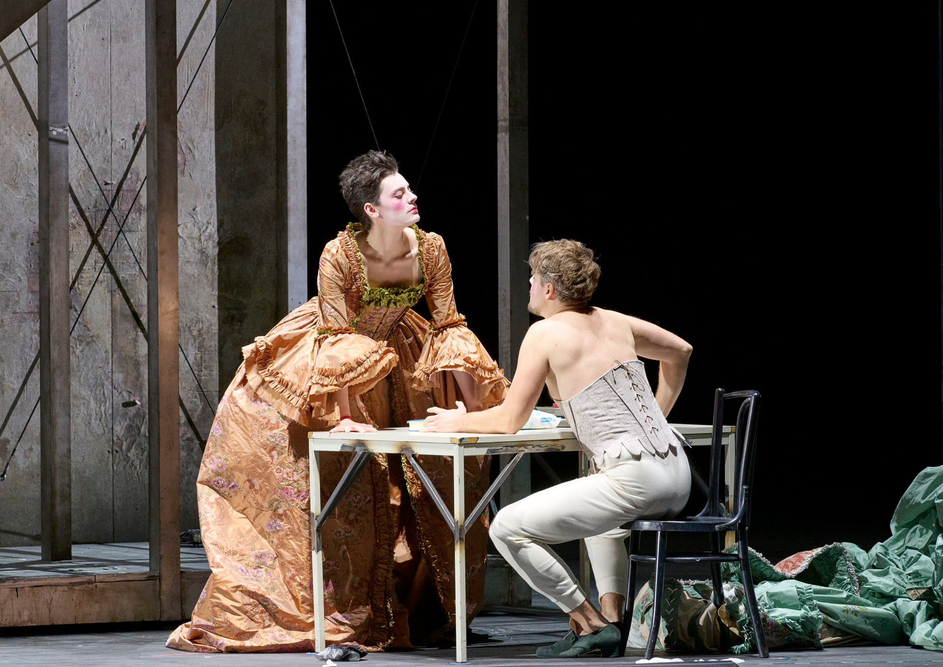 Eine Frau in prächtigem barocken Kleid lehnt sich auf einem Tisch vor und blickt eine Person in Korsett und enger Hose an, die auf einem Stuhl sitzt.