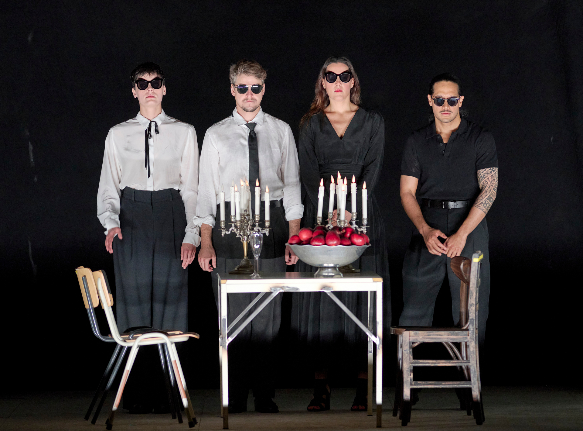 Vier Personen in dunkler Kleidung und Sonnenbrillen stehen vor einem Tisch mit Kerzenleuchtern und einer Schale voller Äpfel auf einer dunklen Bühne.