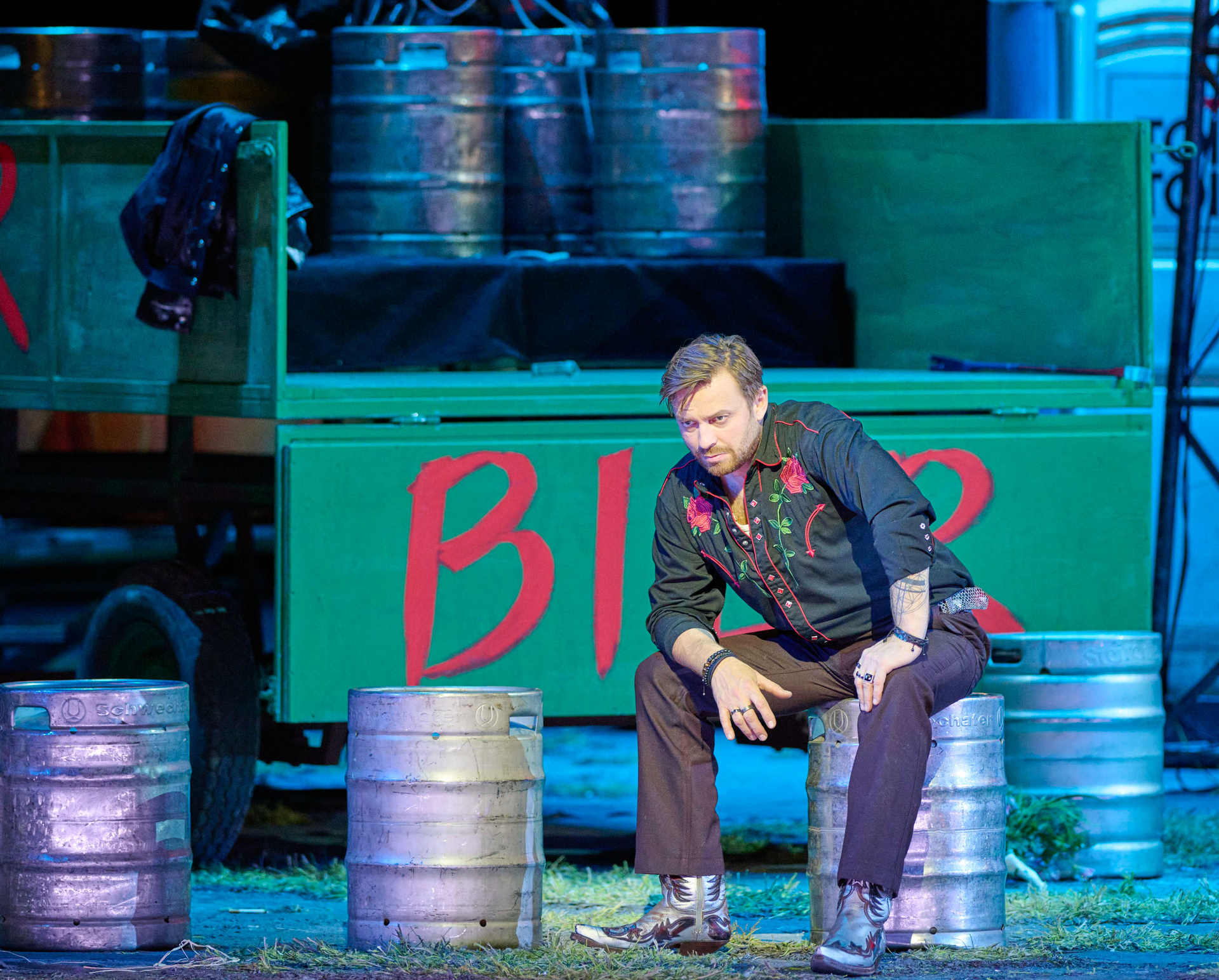 Ein Mann in einem Cowboykostüm sitzt auf einem Metallfass. Daneben und im Hintergrund stehen weitere Fässer.