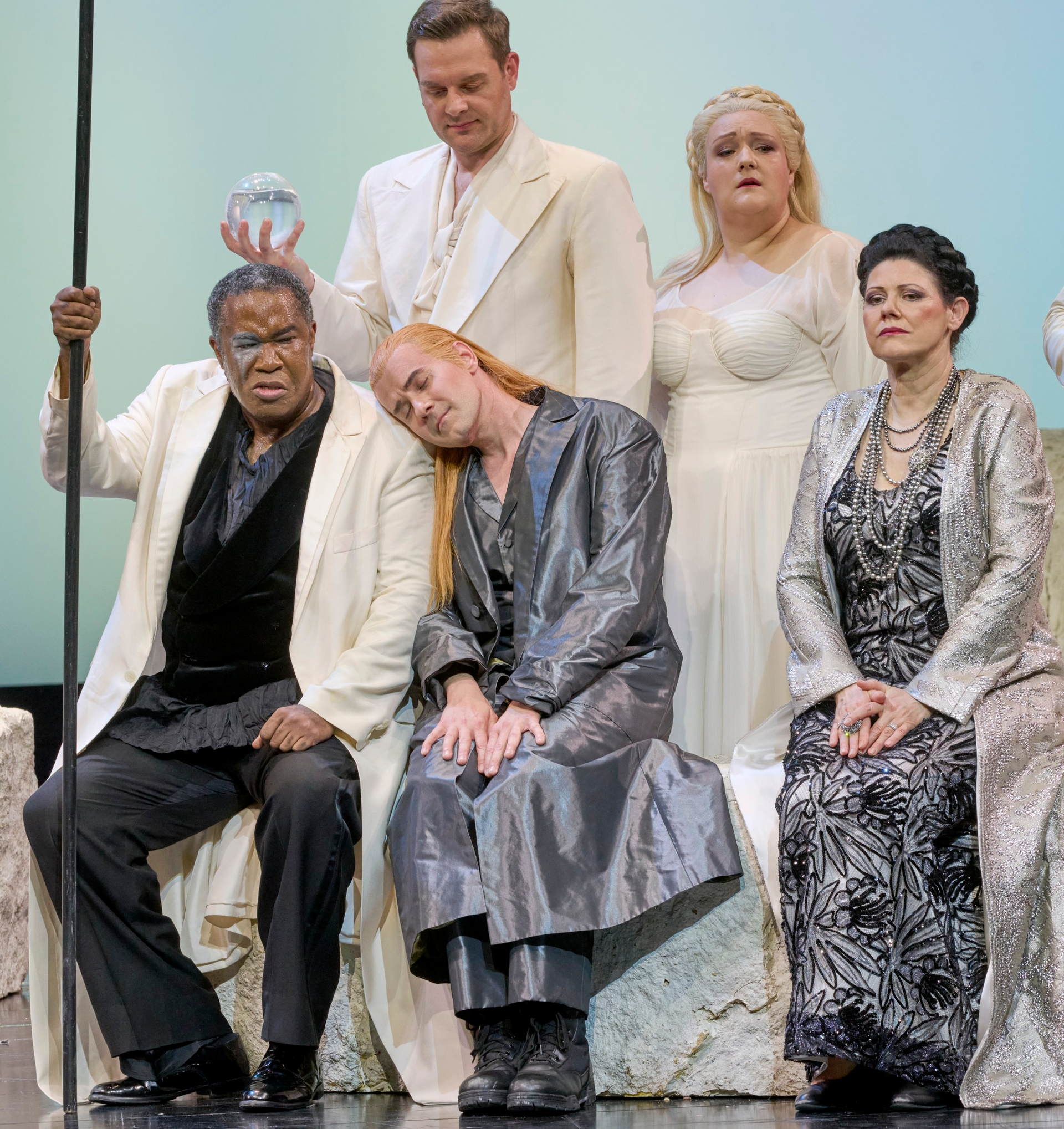 Fünf Personen in festlicher Kleidung sitzen und stehen vor hellem Hintergrund; eine Figur hält einen Stab, eine andere eine Glaskugel.