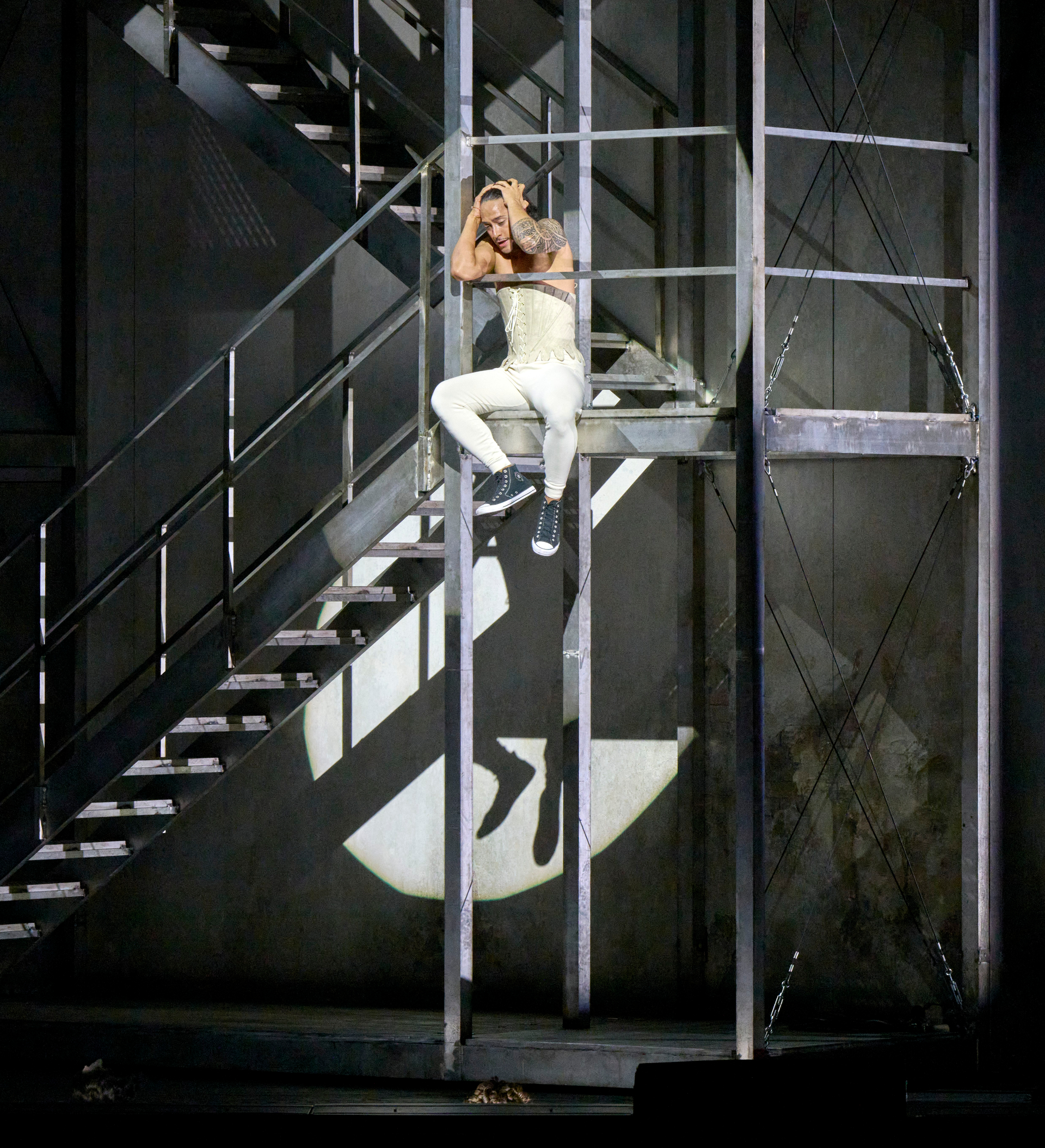 Eine Person in weißem Kostüm hängt an einer Metallstruktur auf der Bühne, hinter ihr ist ein starker Lichtschein mit dem Schatten eines springenden Hirsches zu sehen.