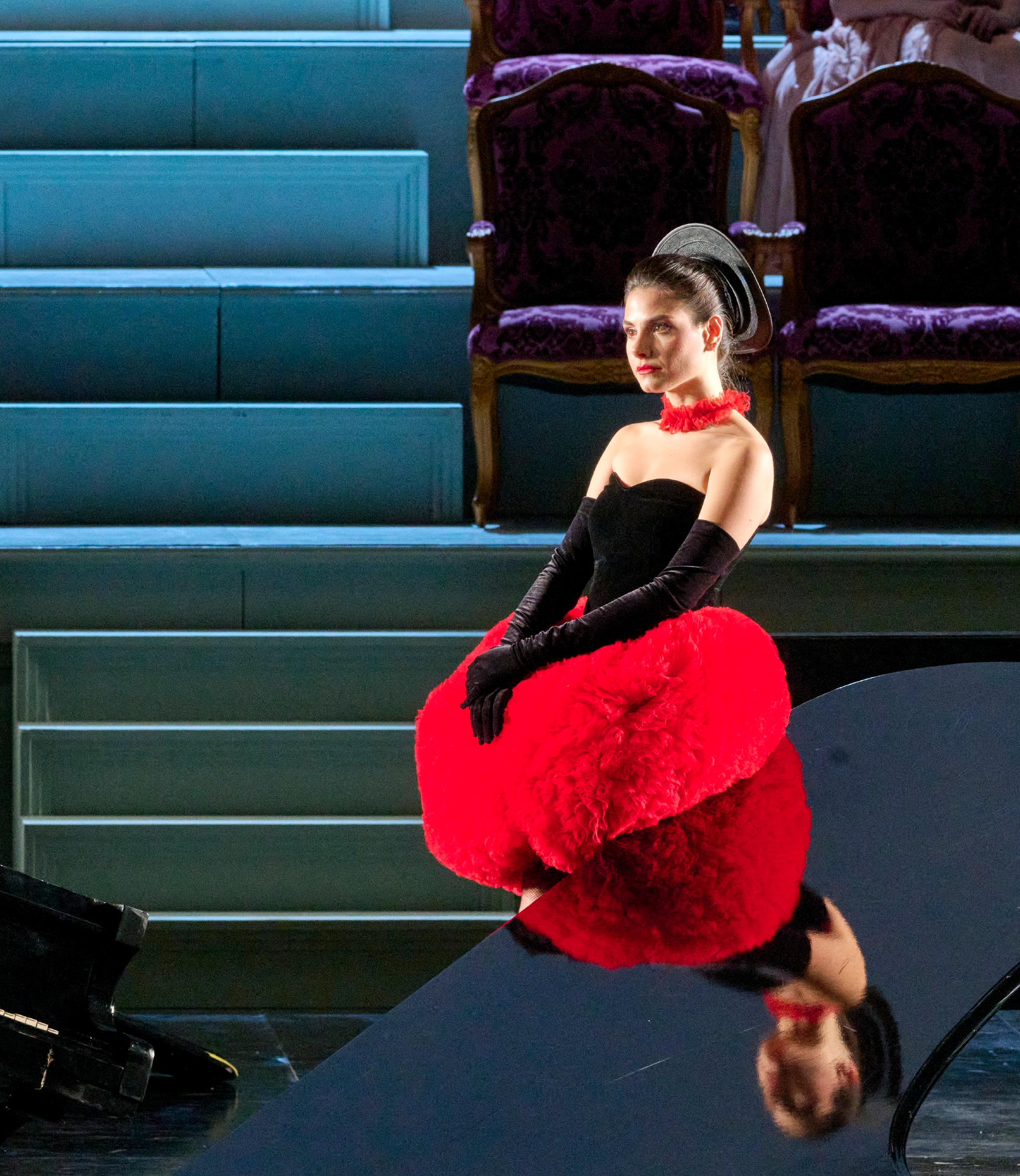 Eine Frau in auffälligem rot-schwarzem Kostüm sitzt lächelnd bei einem schwarzen Flügel, im Hintergrund eine Treppe und leere Theatersessel.