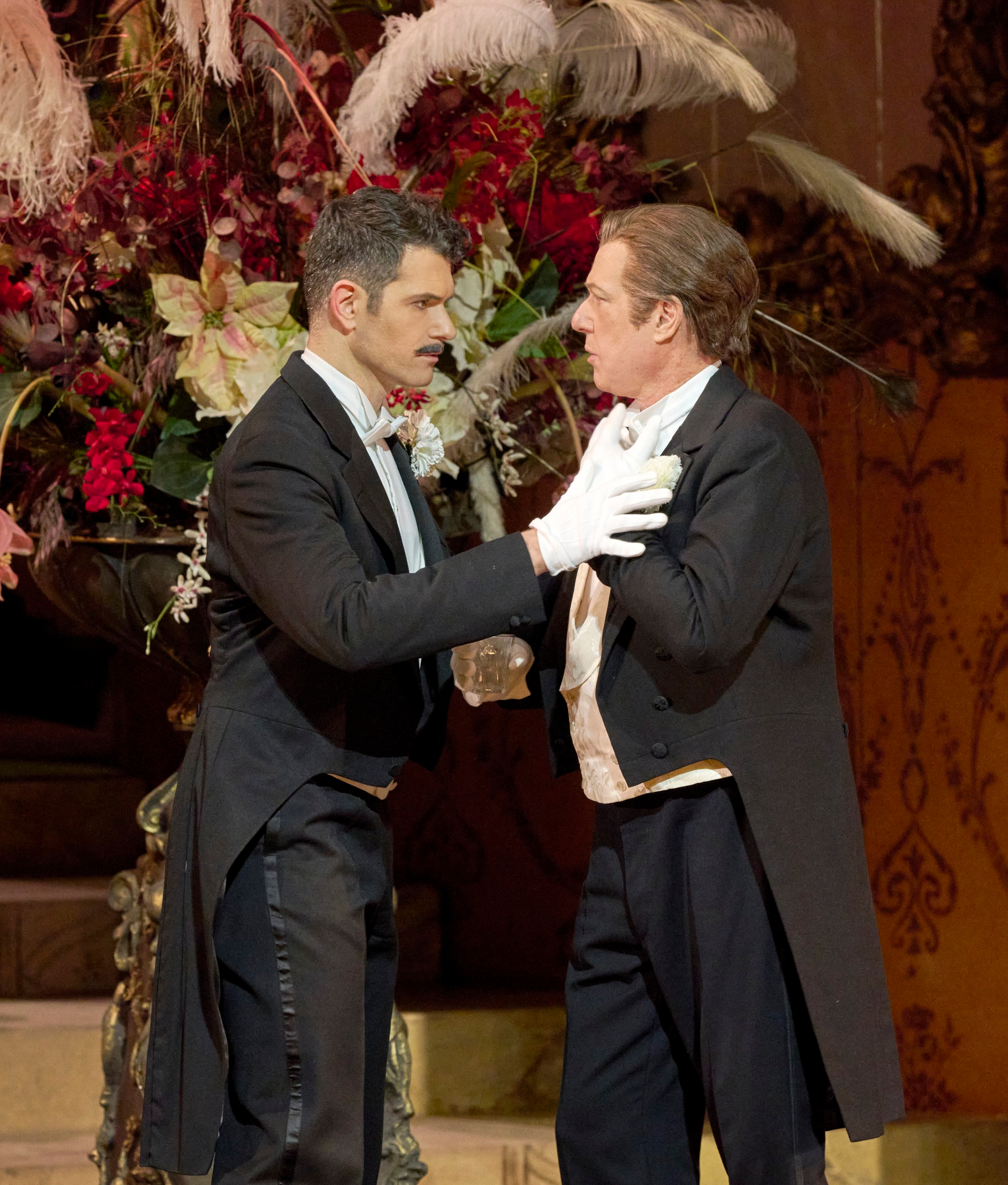 Zwei Männer im Frack stehen sich angespannt gegenüber; einer hält den anderen am Revers fest, im Hintergrund üppiger Blumenschmuck.