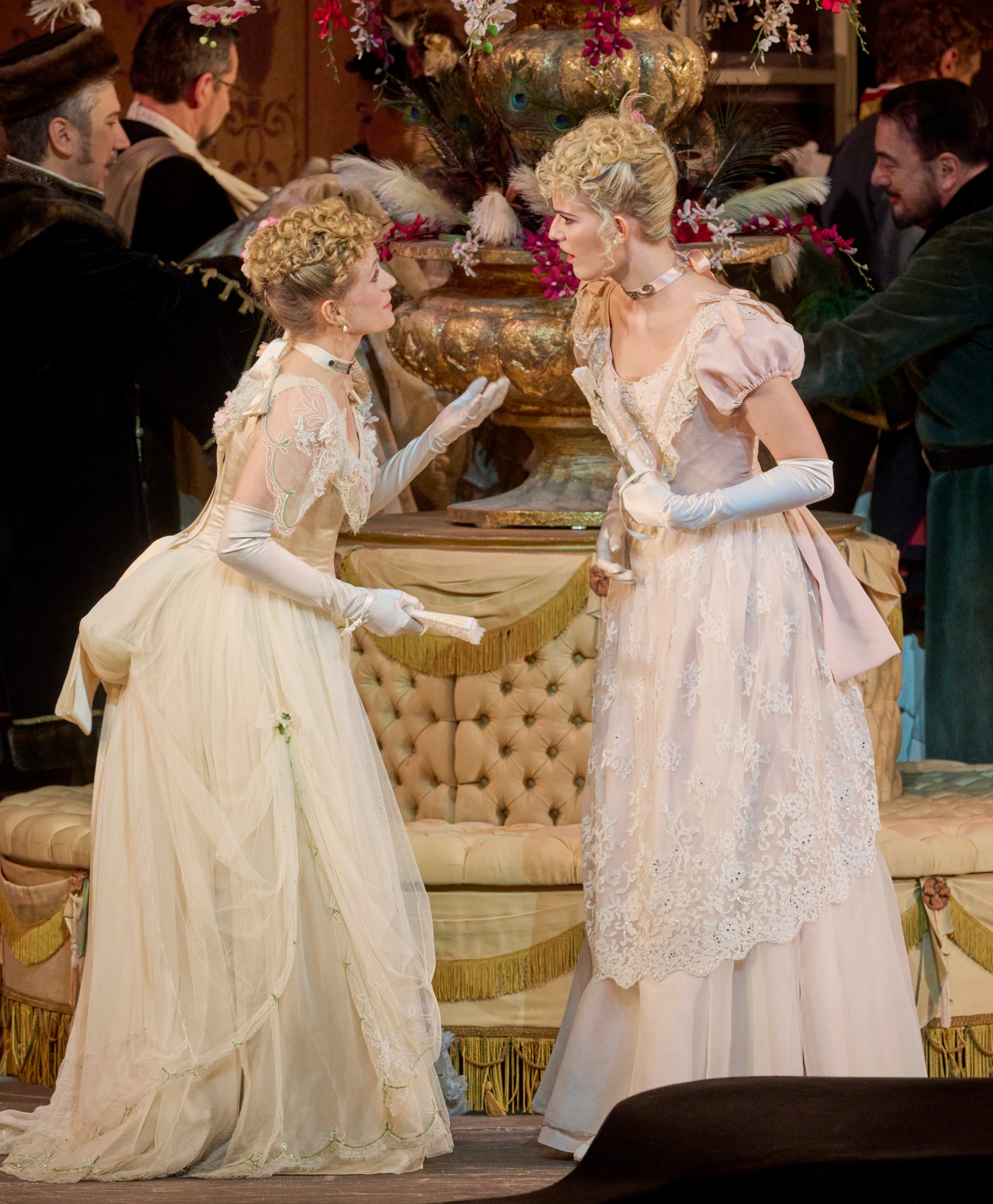 Zwei Frauen in eleganten historischen Kleidern führen eine angeregte Unterhaltung vor einem reich geschmückten Sofa auf einer festlichen Bühne.