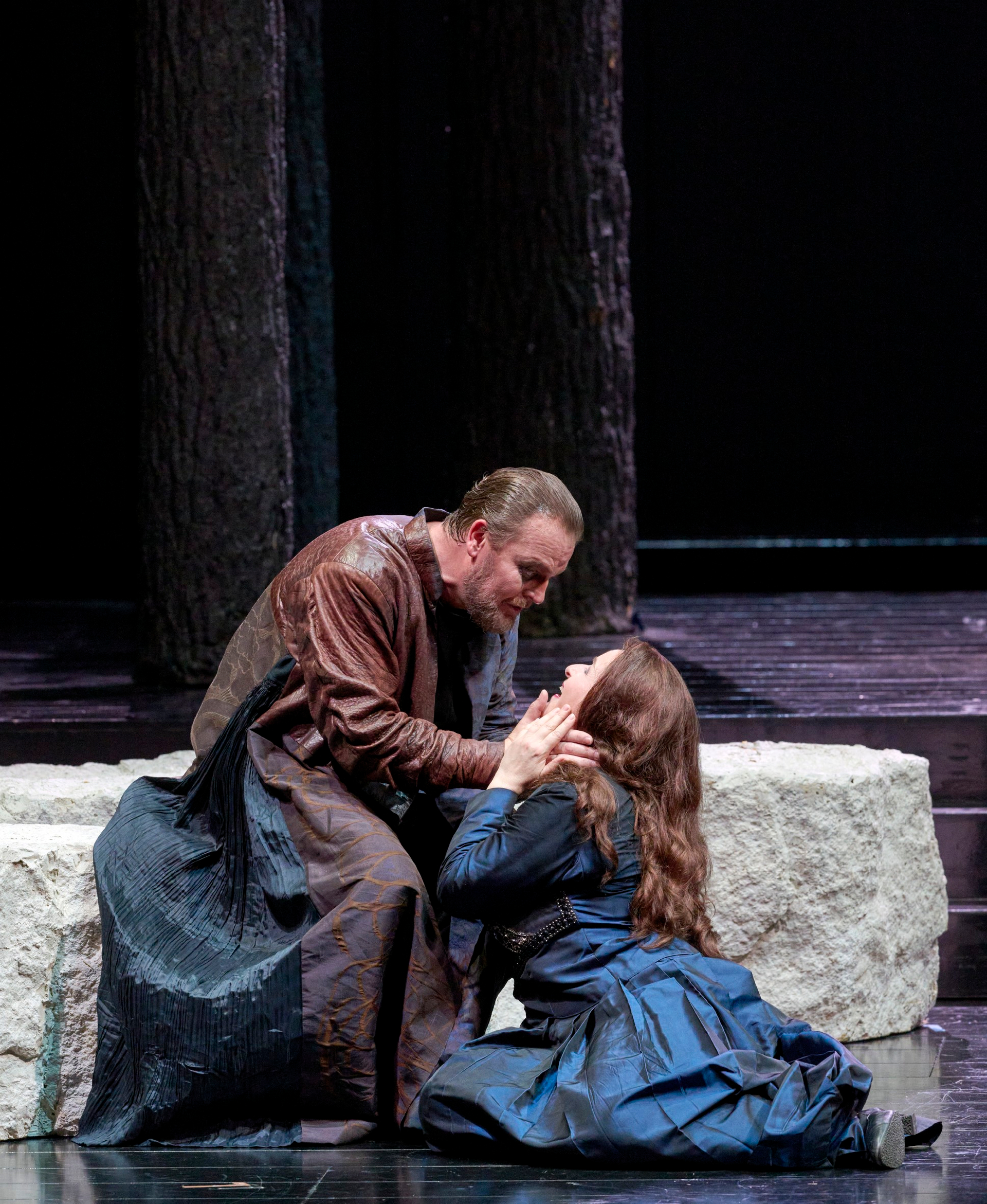 Ein Mann kniet vor einer Frau in dunklem Kleid, hält ihr Gesicht in den Händen und spricht eindringlich zu ihr.