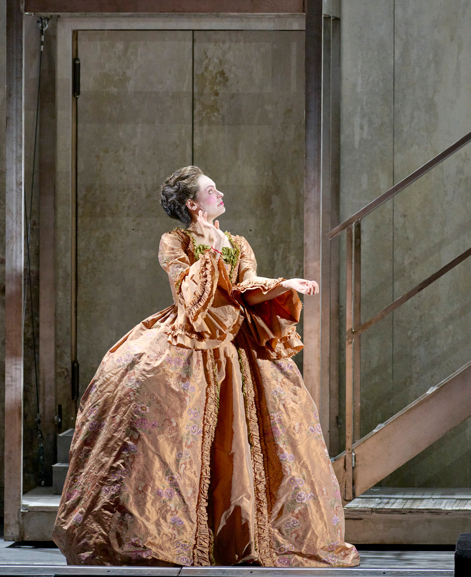 Eine Frau in einem voluminösen, kupferfarbenen barocken Kleid steht vor einer Betonwand und blickt dramatisch nach oben zur rechten Seite.