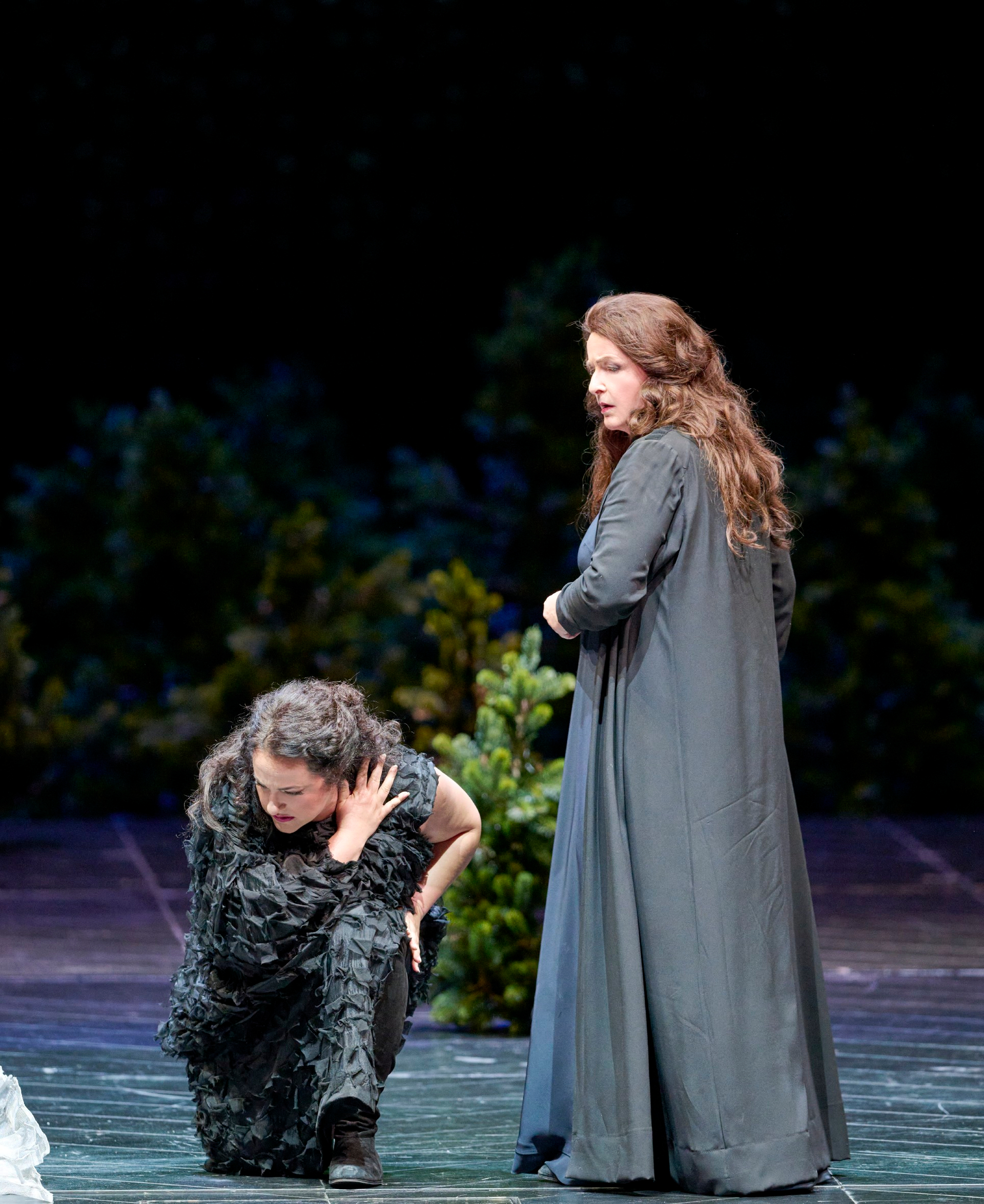 Zwei Frauen stehen auf der Bühne vor einer bewaldeten Kulisse; eine kniet mit gesenktem Kopf, die andere blickt ernsthaft zur Seite.