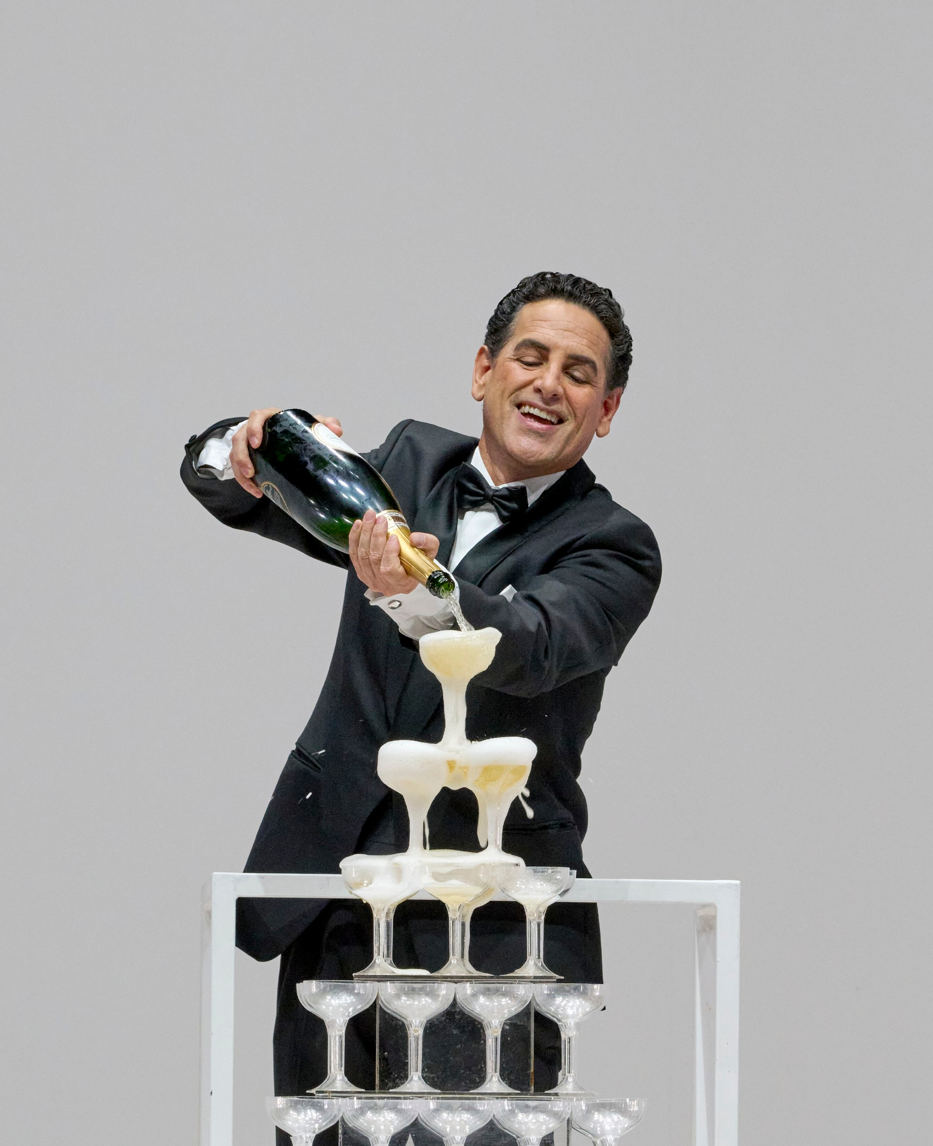 Ein Mann im Smoking lächelt, während er Champagner in eine Pyramide aus Gläsern gießt.