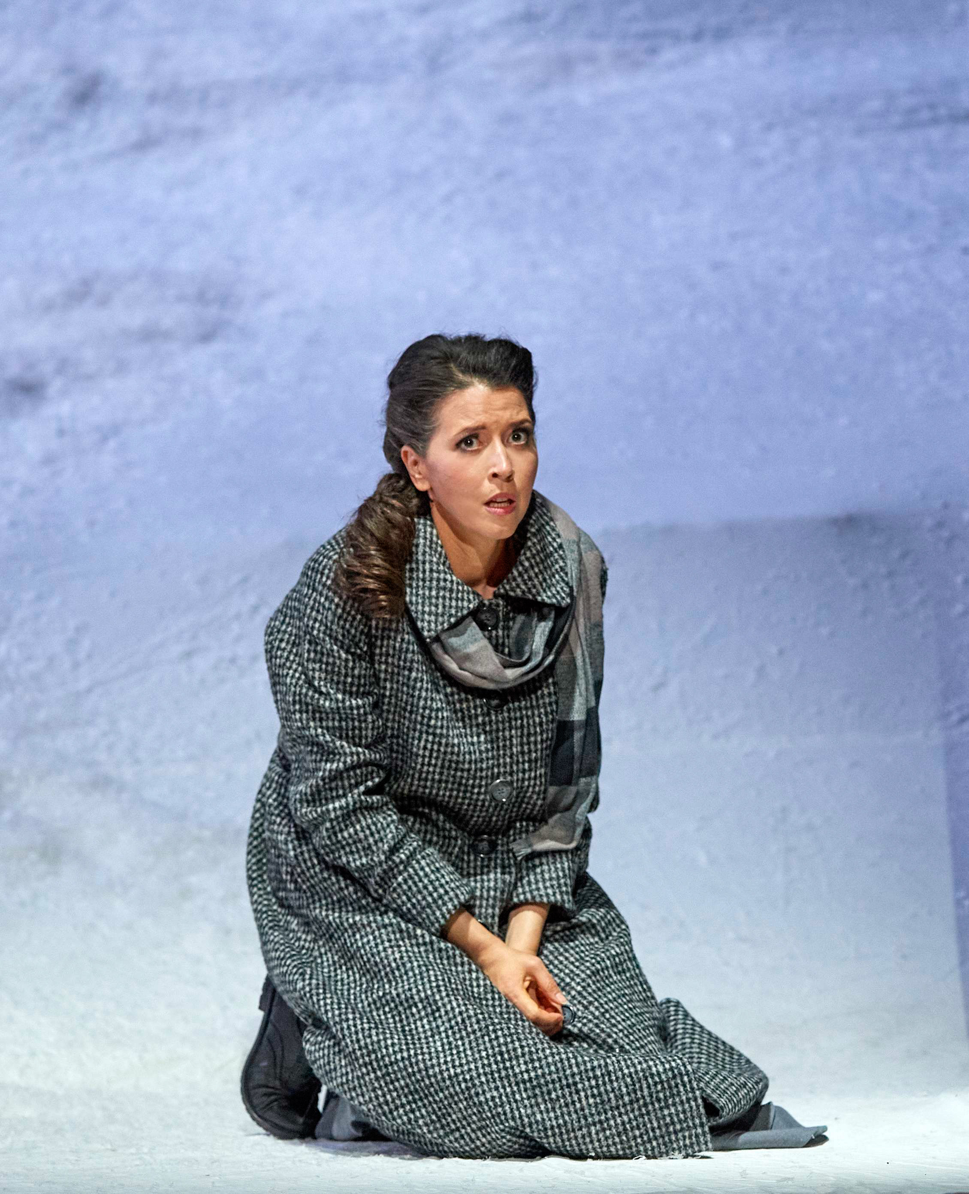 Eine Frau kniet allein auf einer schneebedeckten Bühne und blickt mit ausdrucksvollem, schmerzvollem Gesichtsausdruck in die Ferne.