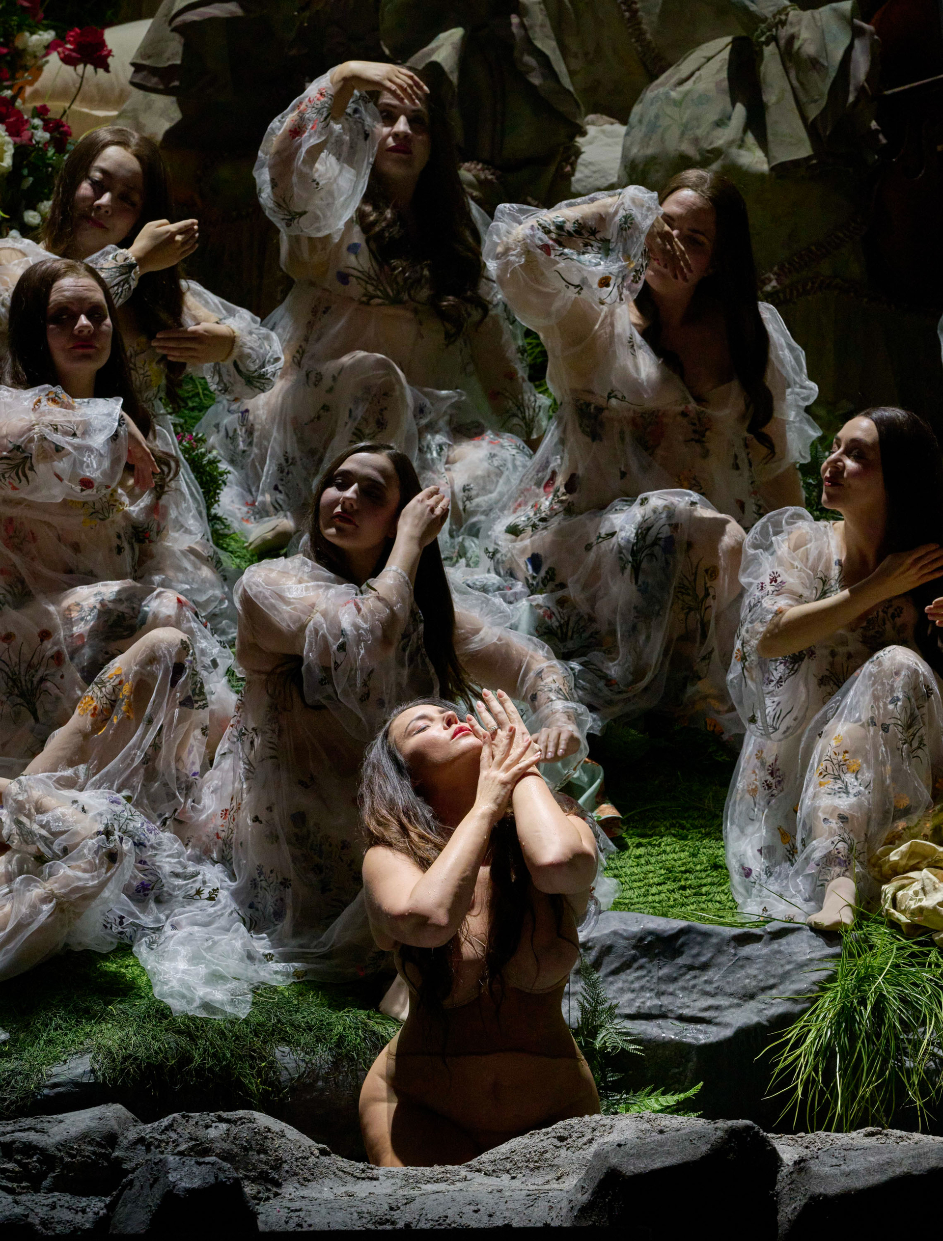 Mehrere Frauen in transparenten, bestickten Gewändern sitzen und knien auf einer grünen Bühne, eine von ihnen hebt emotional die Hände zum Gesicht.