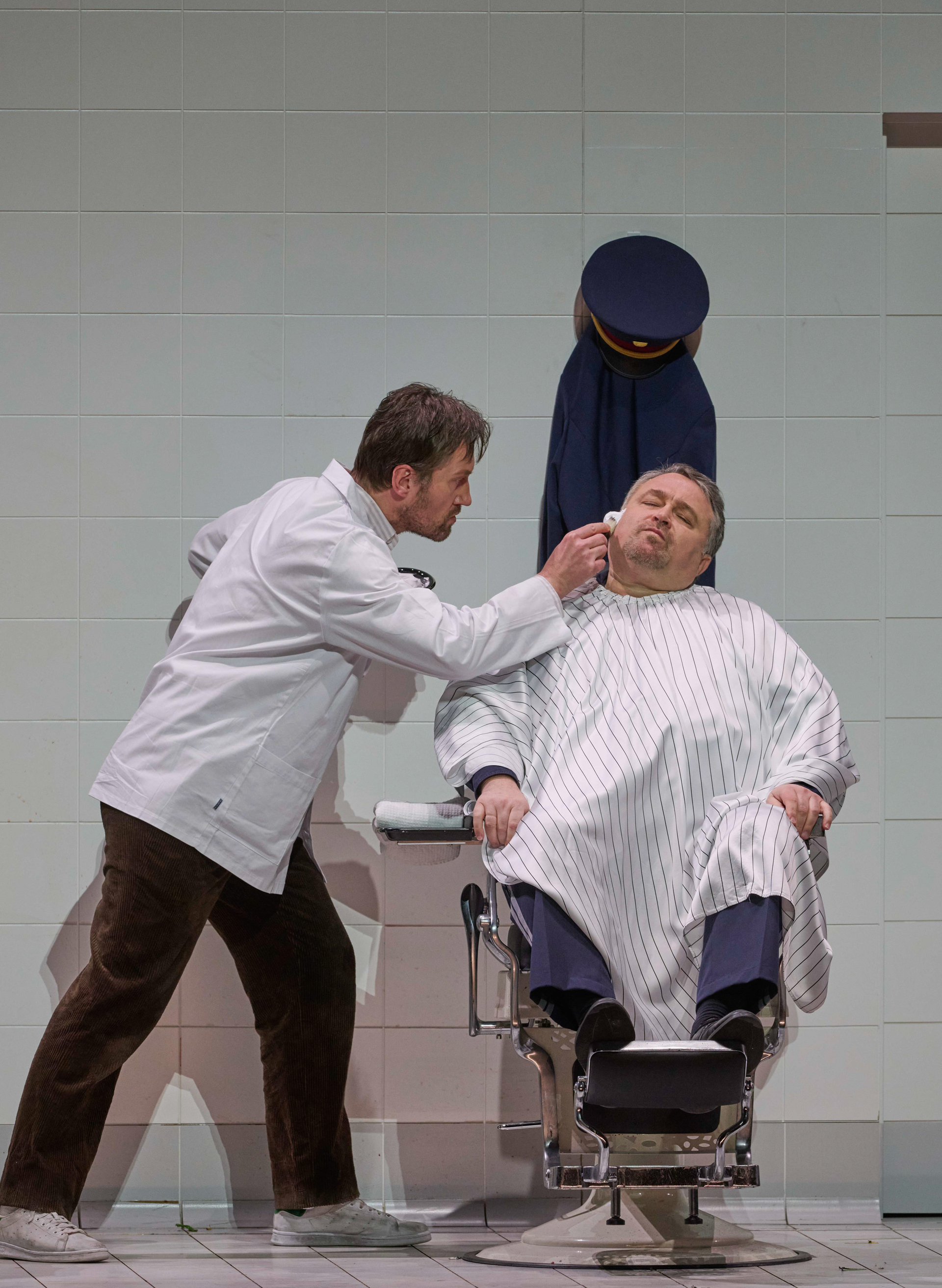 Zwei in weiß gekleidete Männer vor einer gefließten Wand; einer rasiert den Sitzenden im Gesicht.