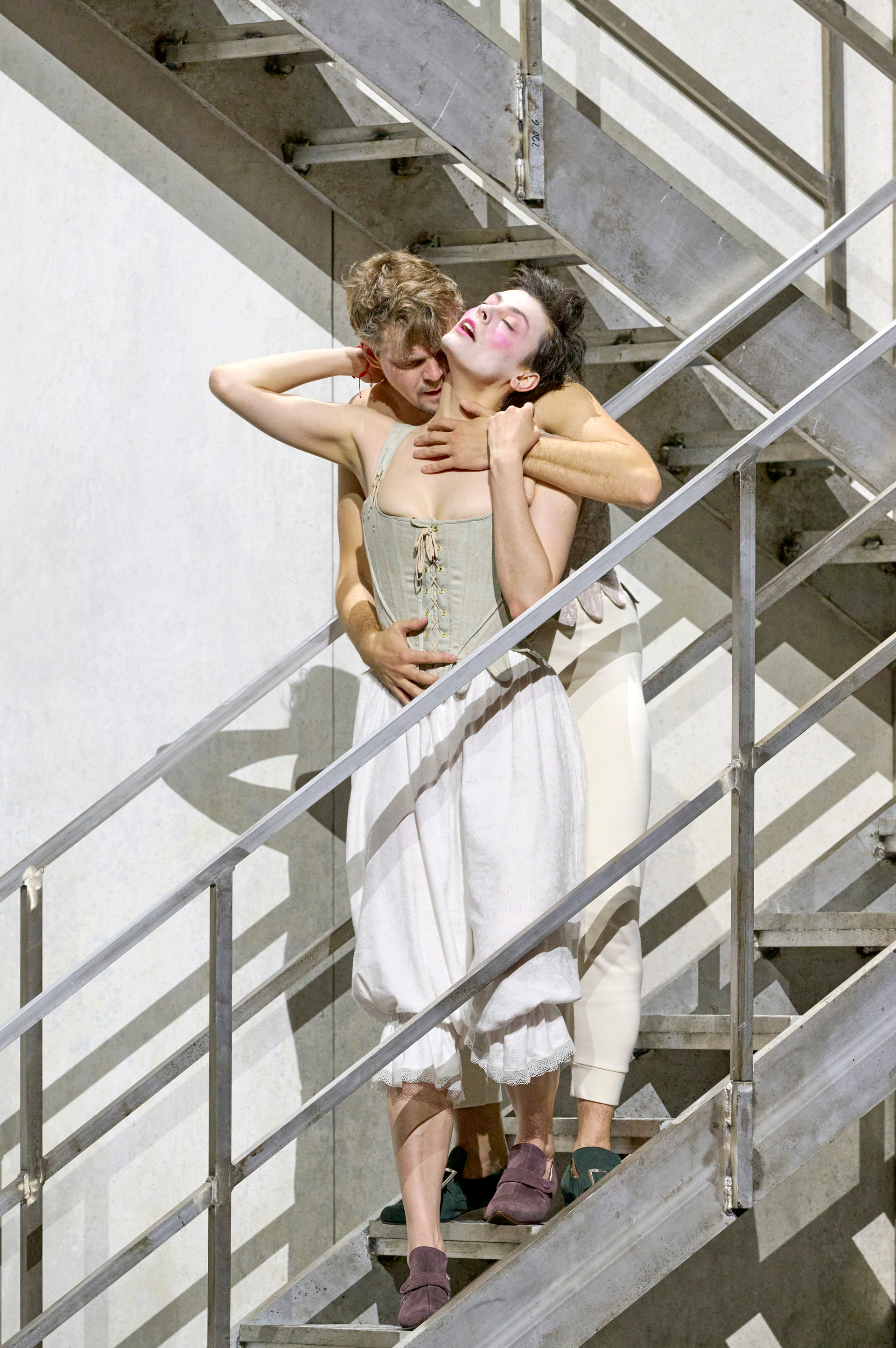 Zwei Personen in historischen Unterkleidern stehen eng umschlungen auf einer Metalltreppe; eine lehnt sich mit geschlossenen Augen zurück in die Arme der anderen.