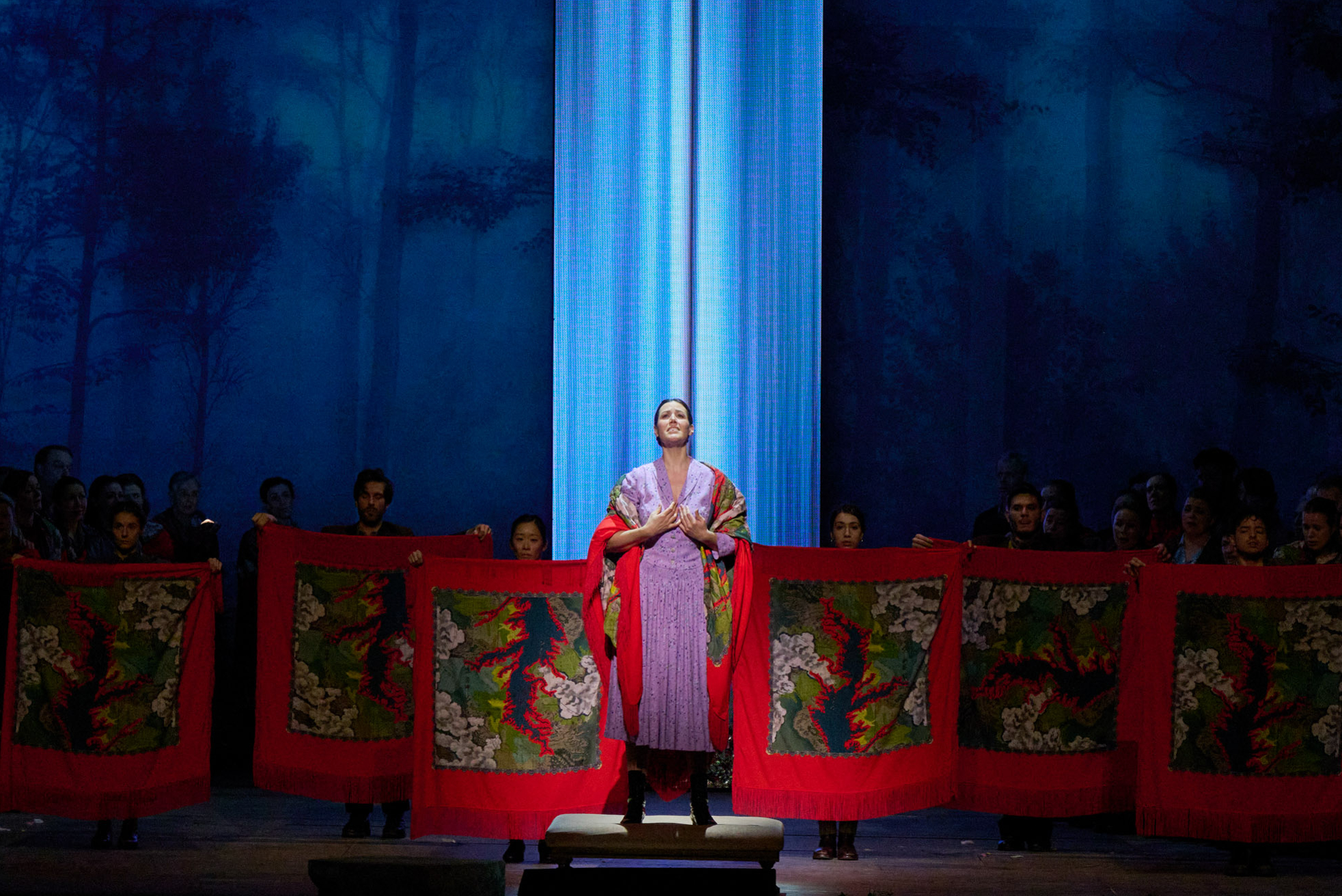 Eine Person im prachtvollen Gewand steht erhöht vor einem Chor, der rote Tücher mit bunten Motiven hält, auf einer Bühne mit Waldkulisse.