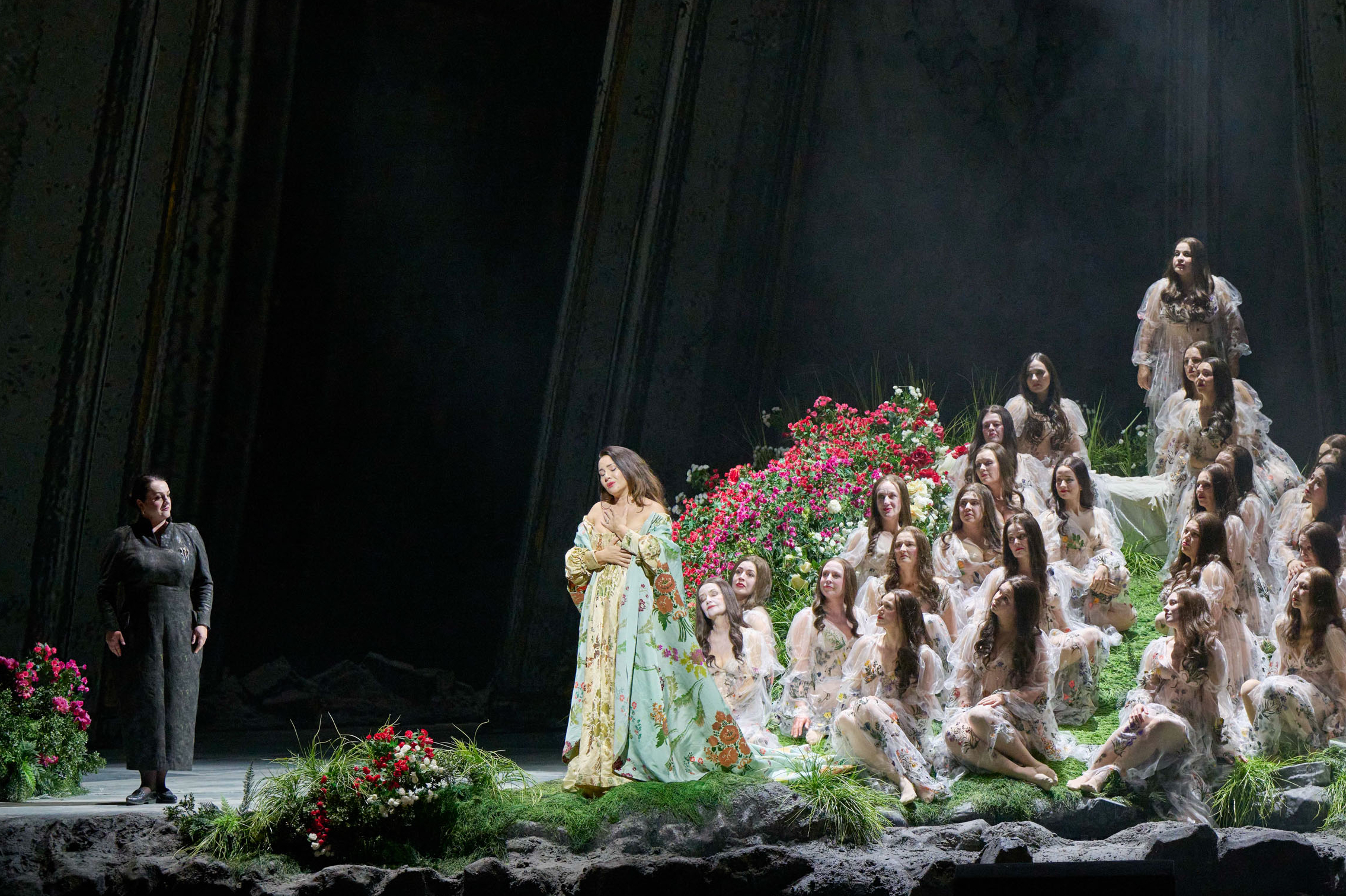 Eine in Gold und Türkis gekleidete Figur steht links auf einer mystischen Blumenbühne, rechts gruppieren sich zahlreiche weiß gekleidete Gestalten auf Terrassen.