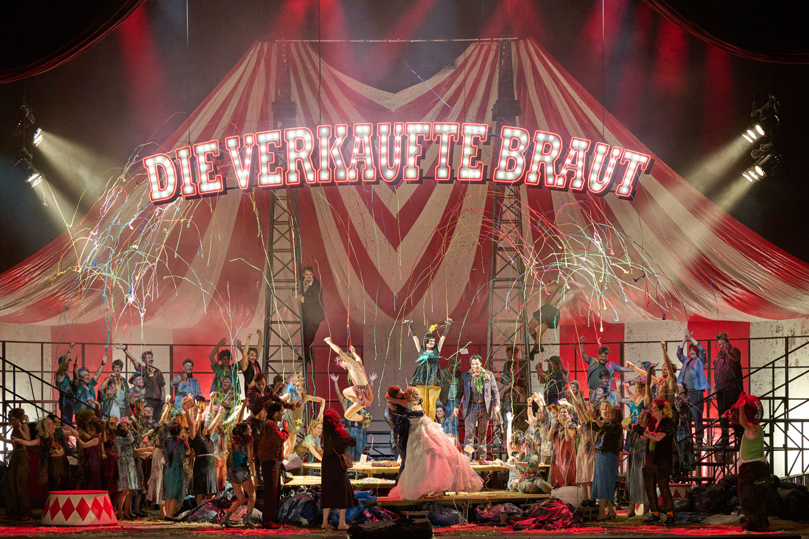 In einem großen Zirkuszelt, an dessen Decke der Schriftzug "Die verkaufte Braut" Hängt, sitzen viele bunt gekleidete Menschen. In der Mitte stehen ein Mann und eine Frau, die sich innig küssen. Von der Decke kommen Luftschlangen.