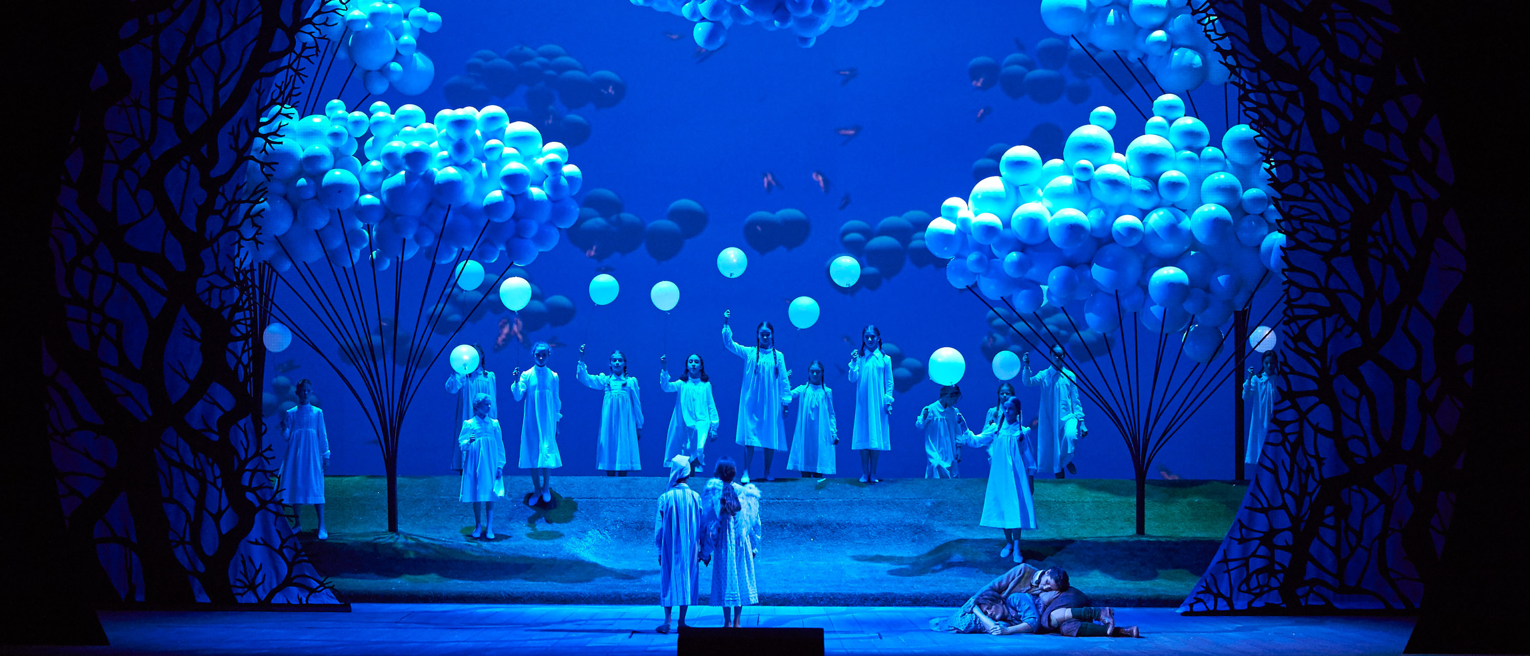 Eine märchenhafte Bühnenlandschaft in Blau mit leuchtenden Bäumen, schwebenden Wolken und Figuren in weißen Gewändern, die feierlich versammelt sind.