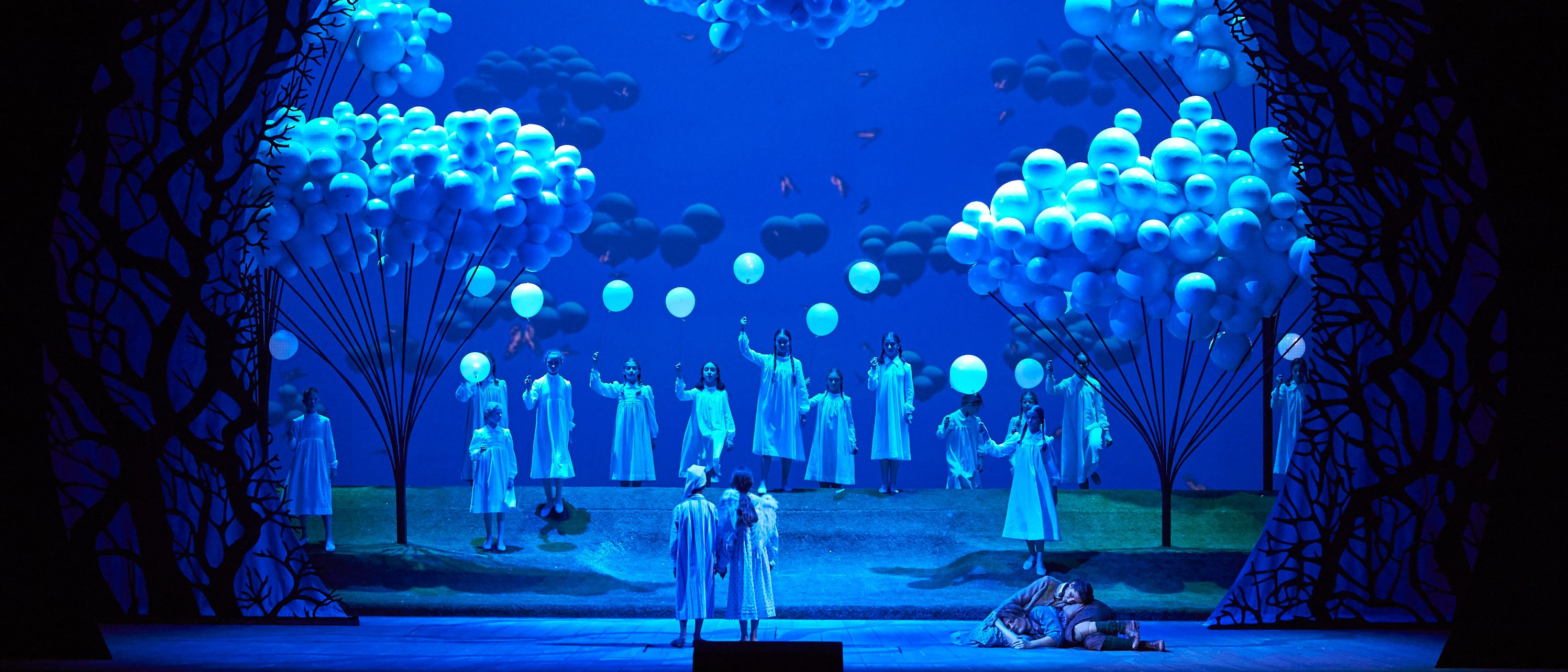 Eine märchenhafte Bühnenlandschaft in Blau mit leuchtenden Bäumen, schwebenden Wolken und Figuren in weißen Gewändern, die feierlich versammelt sind.