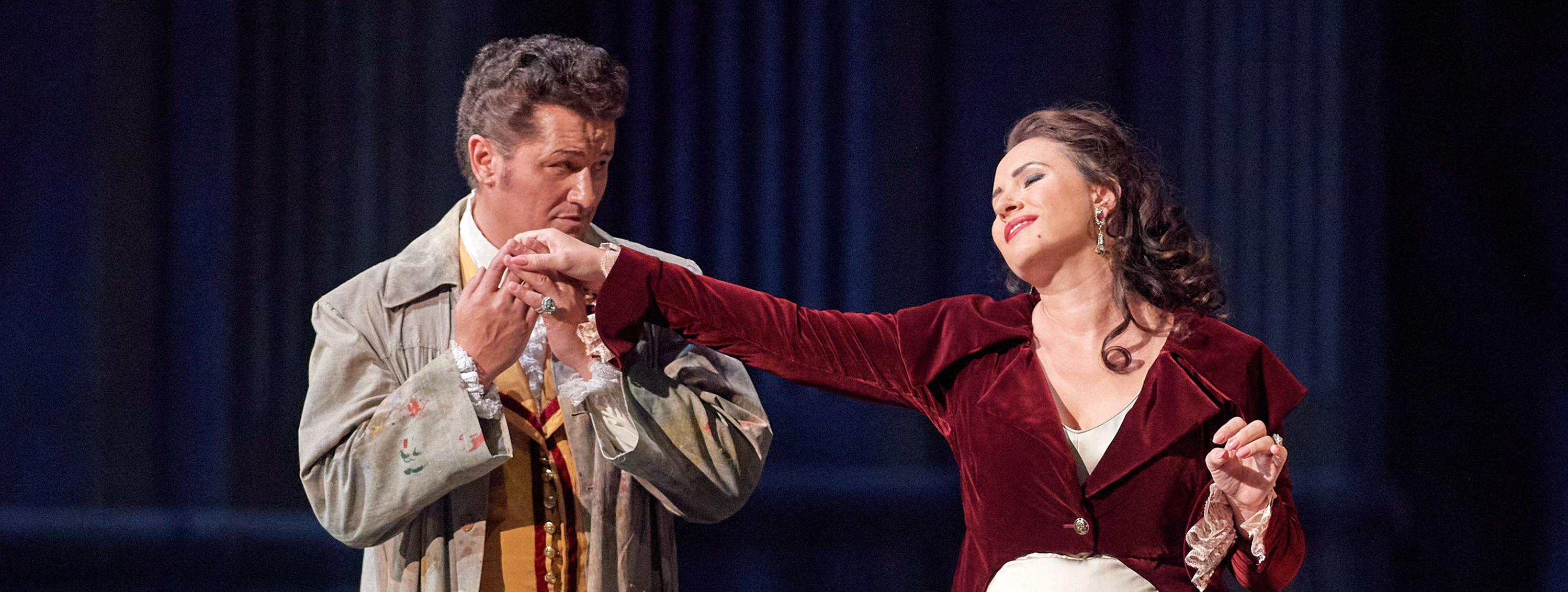 Ein Mann küsst die Hand einer Frau, die in einem roten Samtmantel und einem eleganten weißen Kleid vor ihm steht, während sie ihm liebevoll in die Augen blickt.