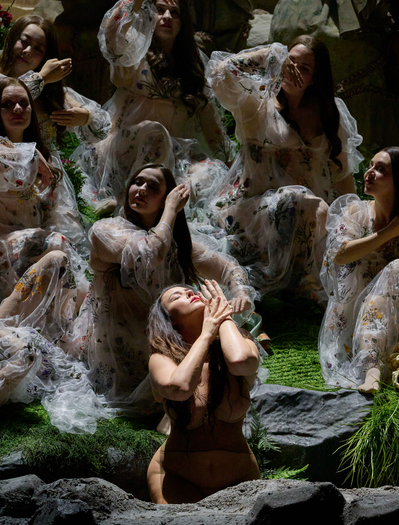 Mehrere Frauen in transparenten, bestickten Gewändern sitzen und knien auf einer grünen Bühne, eine von ihnen hebt emotional die Hände zum Gesicht.