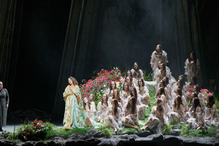 Eine in Gold und Türkis gekleidete Figur steht links auf einer mystischen Blumenbühne, rechts gruppieren sich zahlreiche weiß gekleidete Gestalten auf Terrassen.